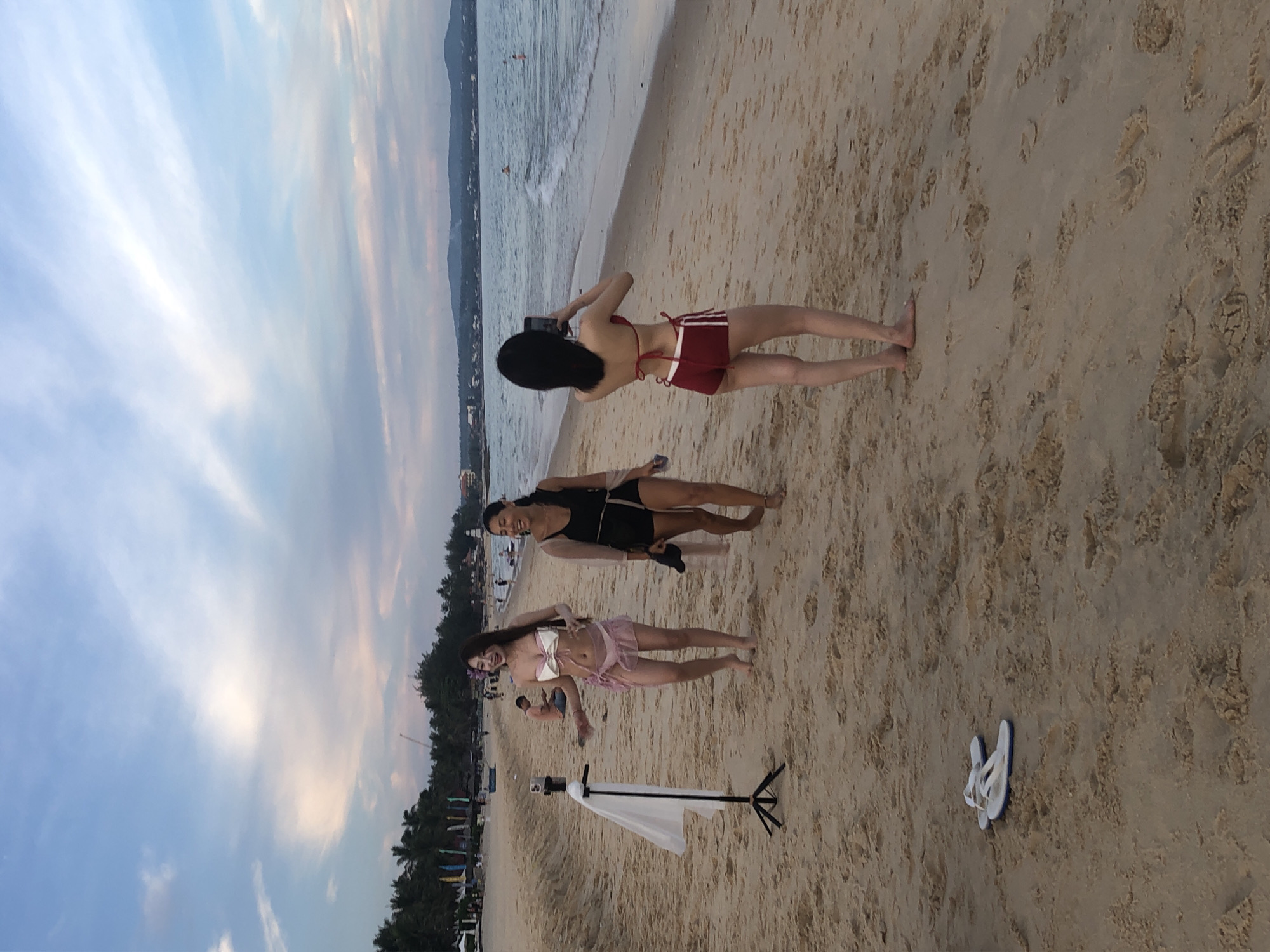 On the sandy beach of Phu Thuy Ward, Vietnam, three young women are present. One woman, wearing a white bikini top and a sheer pink skirt, stands with her arms outstretched as if posing for a photo. To her right, another woman in a black tank top and shorts laughs while holding a light-colored scarf. Further to the right, a third woman, dressed in a red bikini with white stripes, faces away from the camera and holds a smartphone to take a photograph. A portable tripod with a white fabric draped over it is positioned to the left of the first woman. White flip-flops lie on the sand in the foreground. The ocean stretches out behind them, with a hazy coastline visible in the distance under a cloudy sky. The scene appears to be captured during daylight hours.