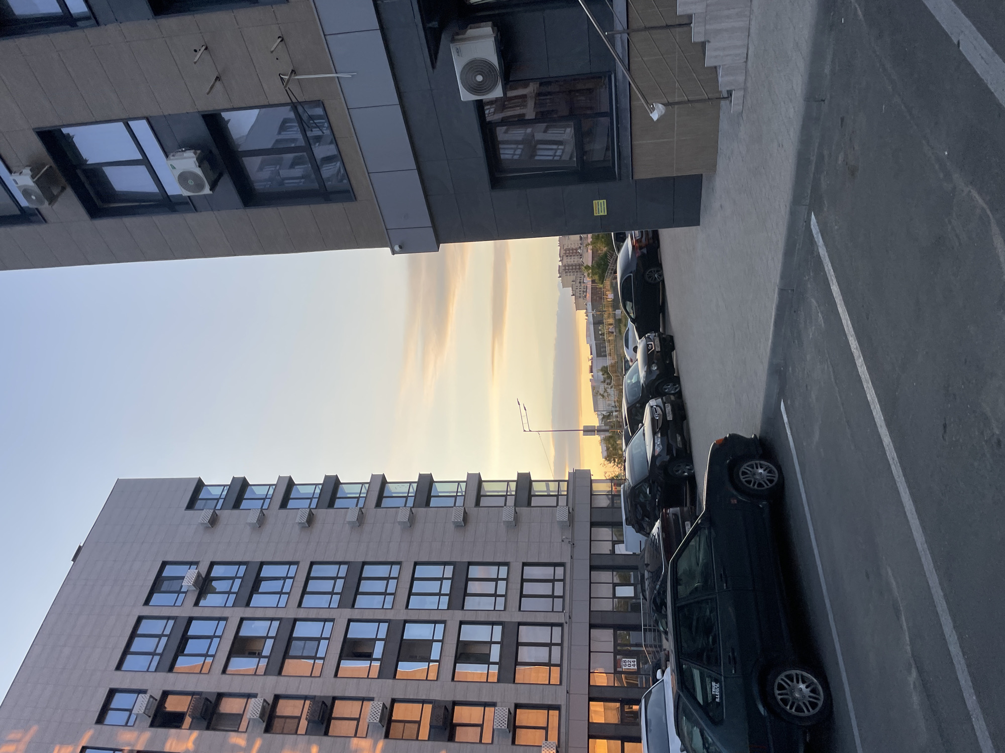 This is a photograph of a parking lot adjacent to a modern, multi-story building with a light-colored facade and numerous windows. The building on the left is taller and appears to be an apartment complex. On the right, another building with a darker facade features external air conditioning units mounted on the walls. Several cars are parked in the lot, including a dark-colored hatchback in the foreground. The sky exhibits the warm hues of sunset or sunrise. In the background, other buildings are visible, suggesting an urban setting. This scene is located in the city of Stavropol, Russia, specifically on Starosel'tsev Street, near house number 68, which is partially visible on the left building.