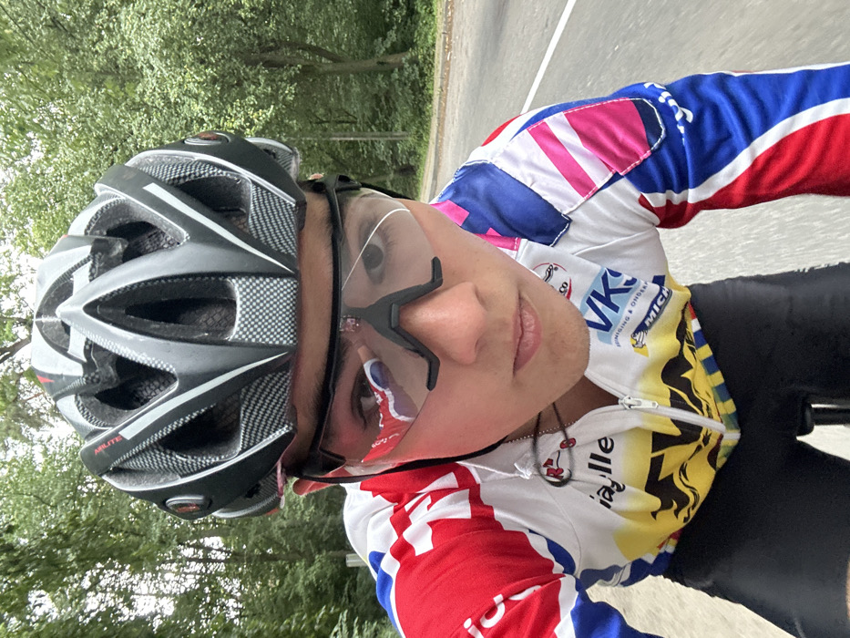 A male individual is depicted in a close-up, first-person perspective, appearing to be engaged in cycling on a paved road. The subject, a young adult, wears a grey and black vented cycling helmet, dark-framed clear cycling glasses with a visible crack on the viewer's left lens and a red reflection on the viewer's right lens. His attire includes a multi-colored cycling jersey, predominantly white, red, and blue, with yellow and black patterns on the lower front. Prominent logos on the jersey include "ia lle", "VKG", "REINIGING & ONDERHOUD", and "MICHELIN". Black cycling shorts are visible below the jersey. A black cord, possibly for earphones, with a small red attachment, is visible around his neck. The subject maintains a neutral expression, looking directly at the camera. The background consists of a grey asphalt road with a white lane line visible on the right, bordered by dense green trees and foliage under natural light, indicative of an outdoor, possibly forested or park-like environment. The scene is located on a road within the city of Bilohorodka, Ukraine.