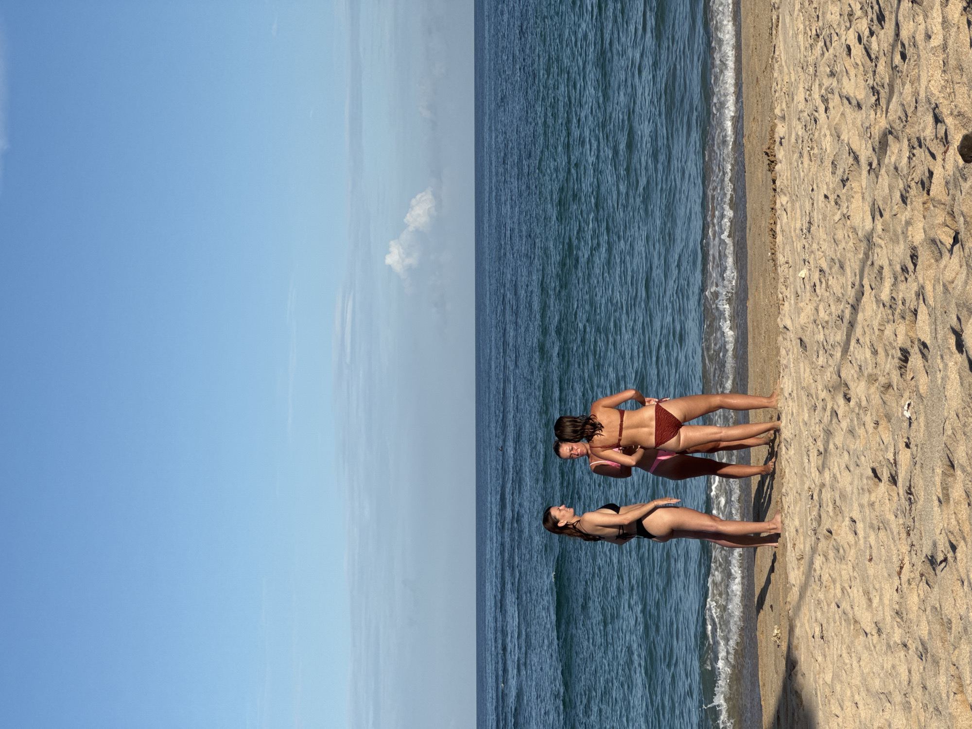 The image shows a beach scene with three women standing near the shoreline. The beach is composed of tan sand. One woman wears a black bikini, while the other two wear brown and pink bikinis. The sea is dark blue and slightly choppy with small waves reaching the shore. The sky is bright blue with some light clouds. The location is likely in Kuta Selatan, Indonesia, given the coastal environment and beach.
