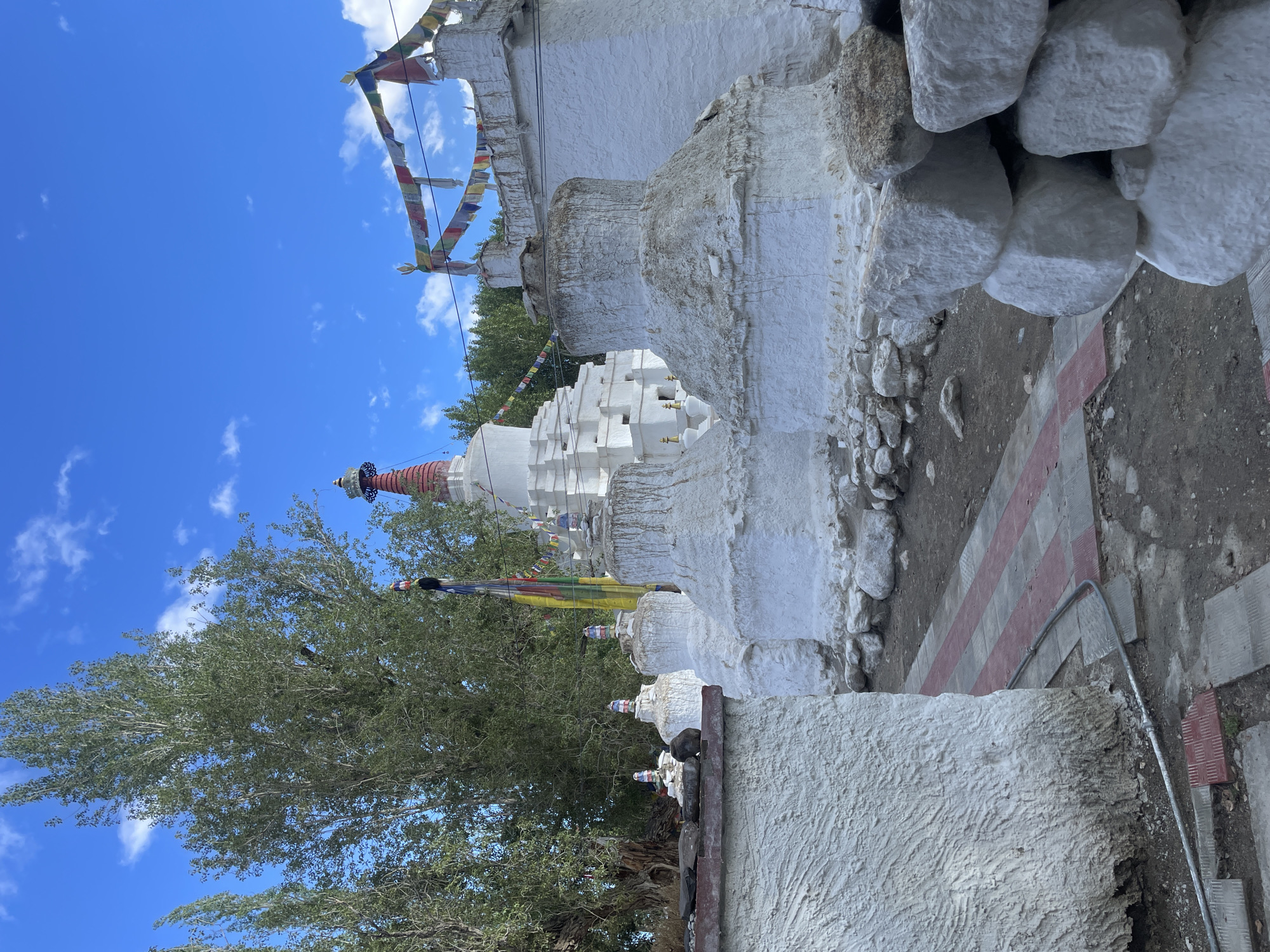 The image shows a Buddhist stupa complex in Leh, India. The stupas are white and appear to be made of plaster over stone. Some of the stupas have prayer flags strung between them. There are trees in the background, and the sky is blue with some clouds. The ground in front of the stupas is a mix of dirt and paving stones. Some of the stupas are partially covered in dirt and debris. The scene appears to be peaceful and serene.