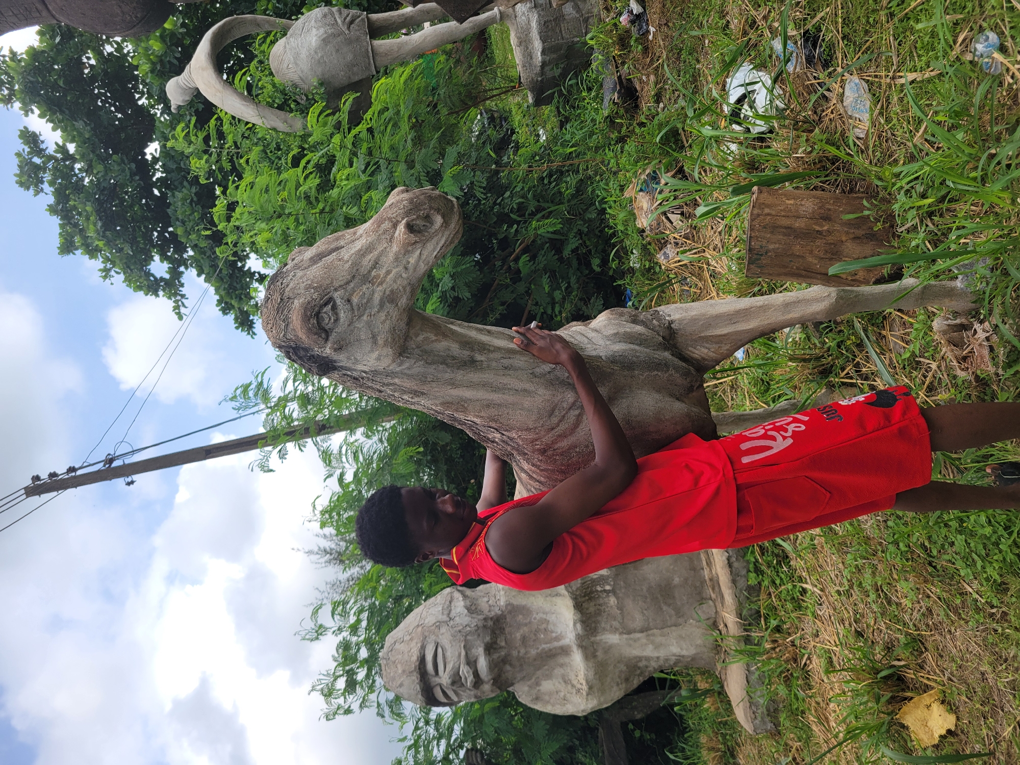 A young Black person wearing a red sleeveless shirt and red shorts stands outdoors, placing a hand on a concrete sculpture of a horse's head and neck. The sculpture is weathered and appears to be part of a larger collection of outdoor art. To the left of the horse sculpture is a large, stone bust of a person with closed eyes and a serene expression. In the background, more concrete sculptures are visible, including a figure of a person from the waist down. The environment is overgrown with green foliage and dry grass, with visible litter on the ground, suggesting an unkempt outdoor space. A utility pole with wires is present in the upper left portion of the image. The sky above is cloudy and blue.