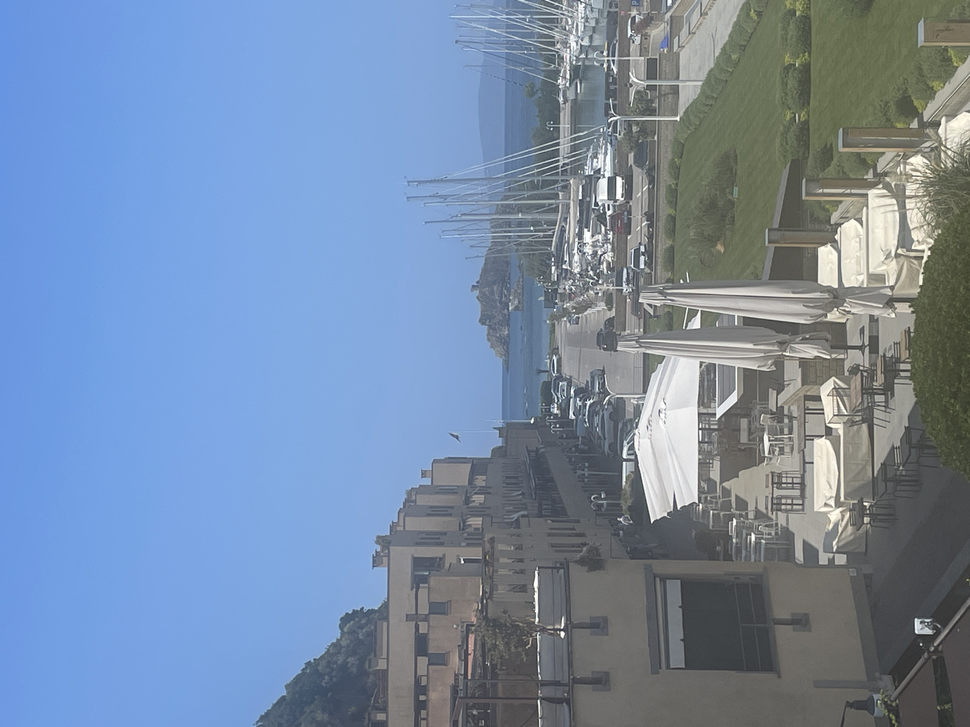 The image shows a sunny day in Punta Ala, Italy. A row of beige buildings lines a street with parked cars. Beyond the buildings, a harbor is filled with sailboats. In the distance, a rocky island with a castle-like structure sits in the sea. A green lawn with manicured bushes borders the harbor. In the foreground, outdoor seating areas with tables and chairs are visible, some covered with protective cloths. The sky is clear blue.