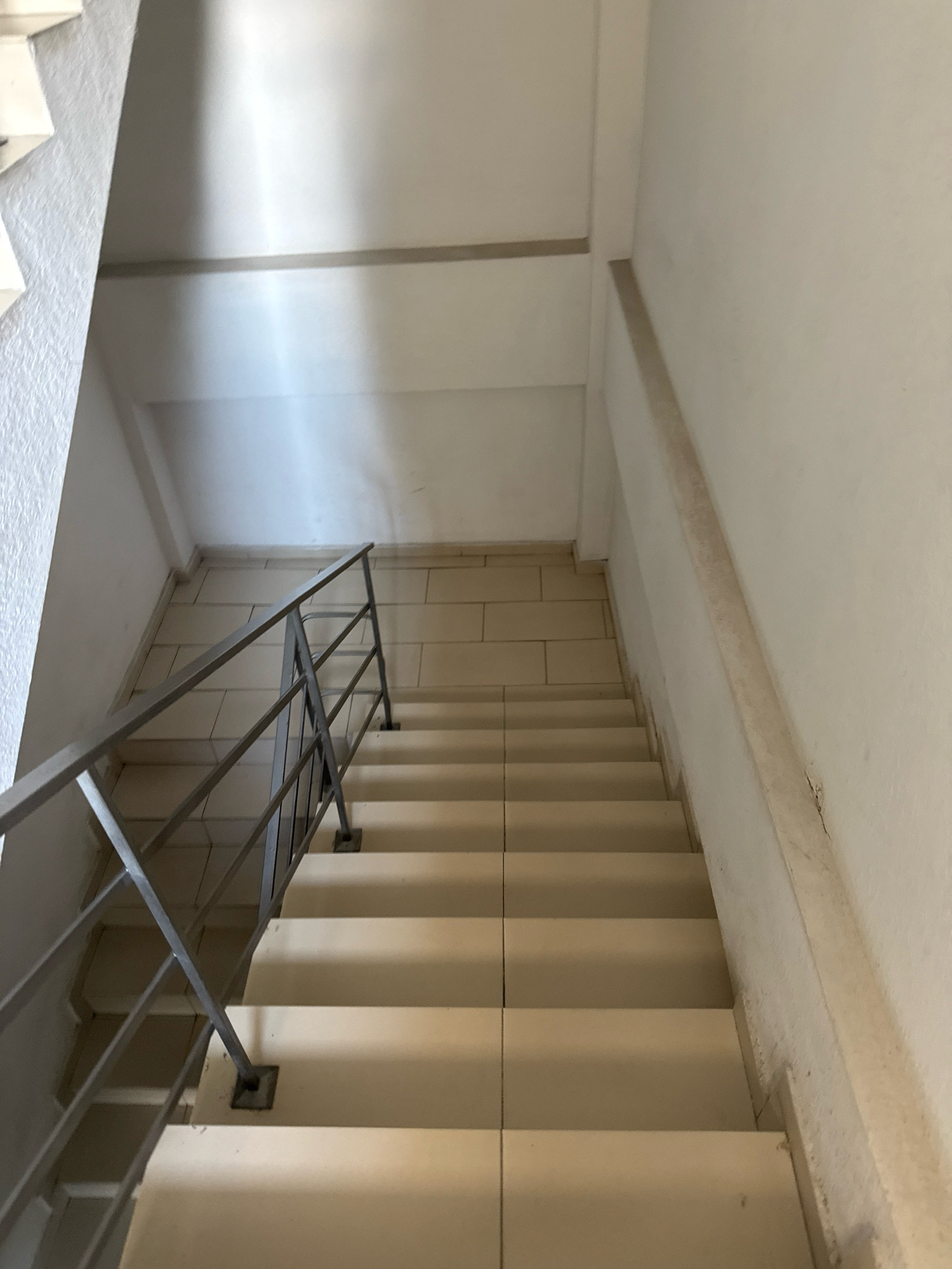An interior view looks down a concrete staircase with light-colored tile treads and risers. A metal handrail with vertical supports runs along the left side of the stairs. The walls are painted white, and the ceiling is also white. The lighting appears to be artificial, casting shadows on the ceiling and walls. The scene suggests a common area within a building, possibly a residential or commercial structure. There are no people or discernible activities depicted in the image. The location of this staircase within Chișinău, Moldova, cannot be determined from the visual information provided.