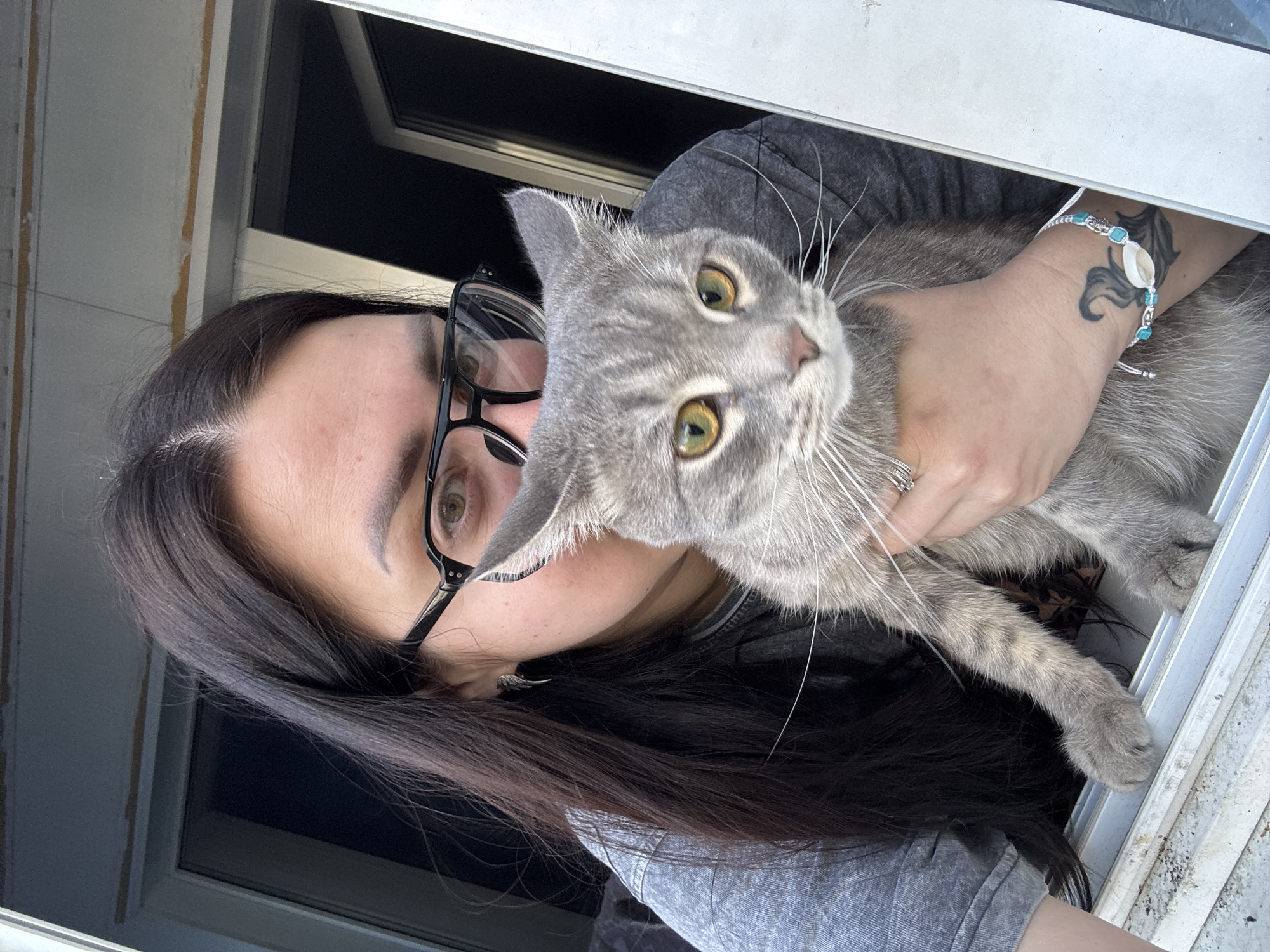 An image, rotated 90 degrees counter-clockwise, features a person holding a gray tabby cat. The person, positioned on the left side of the frame, has long dark hair, dark-framed glasses, and appears to be wearing a dark t-shirt or sweatshirt. Their left eye, visible through the glasses, is a light brown or hazel color. Notable details include a tattoo on their left forearm, resembling a tribal or stylized wave design, and a bracelet with light blue beads and a white shell charm worn on the same wrist. Their expression is neutral, directed towards the camera.
The cat, held securely by the person's hands, is prominently featured in the center and right of the frame. It has distinctive gray fur with faint tabby stripes and large, expressive yellow-green eyes with black pupils. Its white whiskers are long and prominent. The cat's gaze is also directed towards the viewer, appearing alert and curious.
The background is a mix of indoor and possibly outdoor elements. A white window or door frame runs horizontally across the top and bottom of the rotated image. Behind the subjects, a dark, rectangular area suggests a window looking into a darker space or another room. Portions of a lighter-colored wall or paneling are visible near the top left. The lighting suggests it is daytime. There is no visible text in the image.