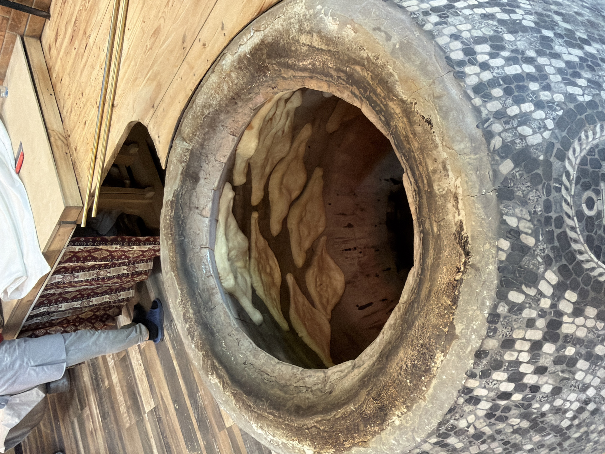 The image, rotated 90 degrees clockwise, depicts an indoor scene focused on traditional bread baking. A large, cylindrical tonir oven dominates the right side, its wide opening revealing several pieces of light-colored dough, shaped like elongated ovals or teardrops, adhering to the hot inner walls as they bake. The oven's rim is dark and worn from heat and use.
The exterior of the tonir is decorated with an intricate mosaic pattern of alternating dark and light grey/white small tiles, resembling a checkered or scale-like design. A decorative band, possibly with a stylized leaf pattern, is also visible within the mosaic.
To the left, a person is partially visible, wearing light grey pants and dark blue slip-on shoes, standing on a wooden plank floor. A patterned rug, primarily red with geometric designs, lies nearby. In the background, a white garment hangs, and several long, thin, golden-colored metal rods, likely tools for handling bread in the oven, rest on a wooden surface. An arched wooden structure is also seen further back.
The setting suggests a traditional bakery or kitchen, with bread baking as the central activity. The presence of the tonir oven and the method of baking flatbreads are characteristic of Armenian culinary traditions, consistent with the location context of Tsovagyugh, Armenia. No text is visible in the image.