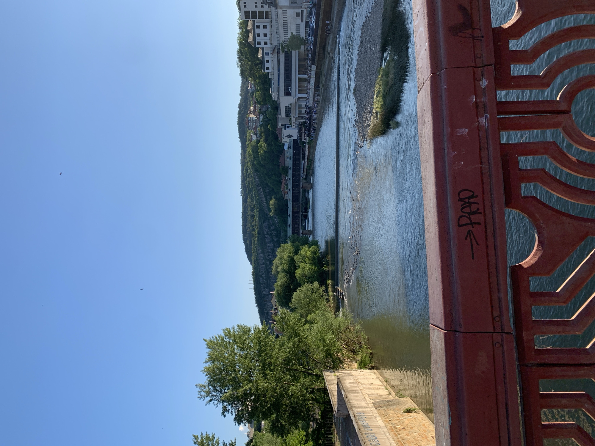 The image depicts a cityscape taken from a bridge. The scene features a river flowing beneath the bridge with a small island covered in vegetation in the middle. The far bank is lined with buildings that rise up the hillside, with trees interspersed. The bridge's railing is in the foreground, painted in red, and bearing some graffiti. The sky is clear blue. Location is the city of Unknown, Unknown.