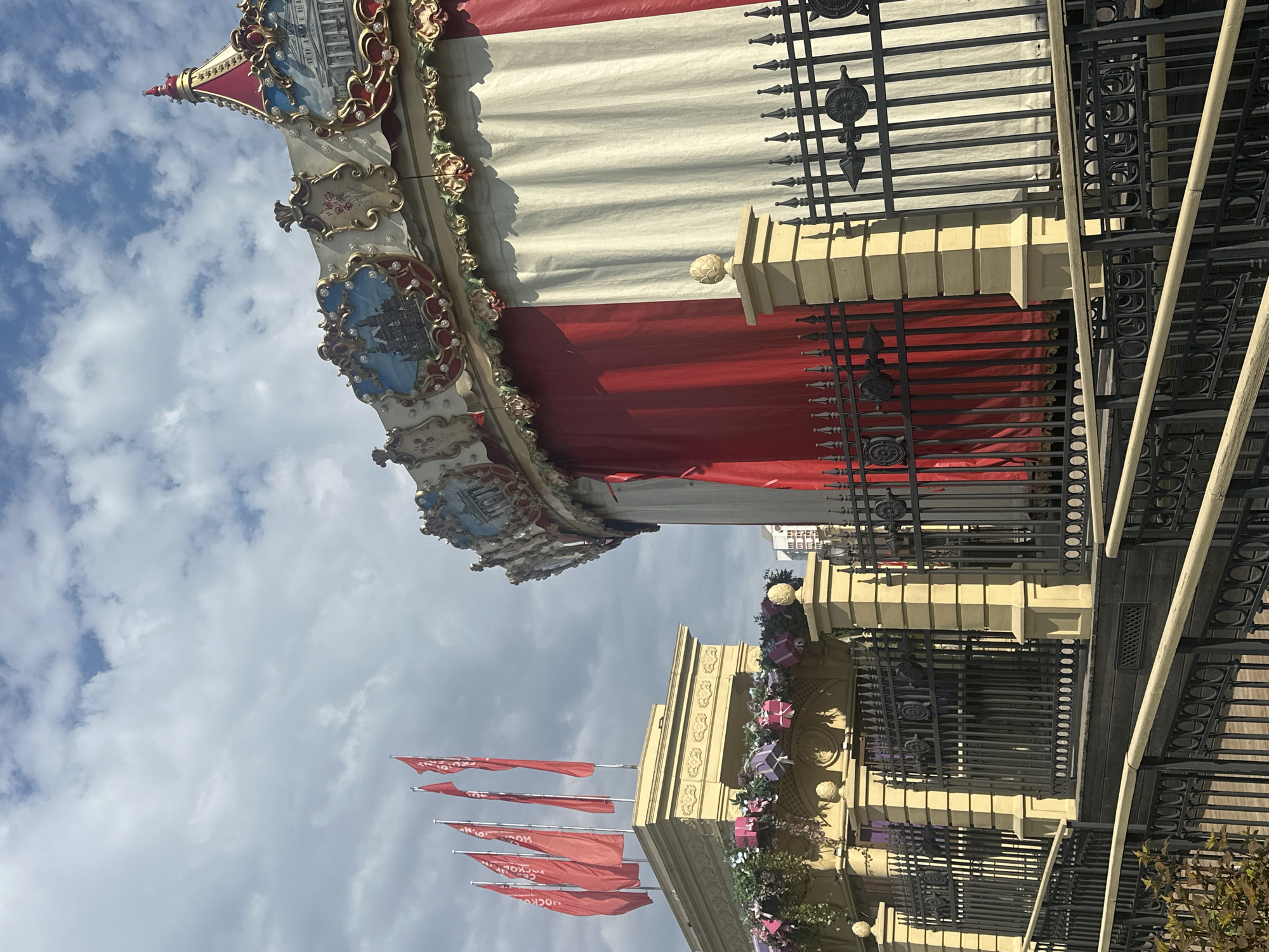 The image shows a portion of a carousel's decorative roof with ornate trim and painted scenes, positioned against a backdrop of a cloudy sky. Below the carousel roof are building elements, including a beige facade with decorative floral arrangements and wrapped gift decorations. A black wrought-iron fence is positioned alongside the building. Several red flags are on flagpoles near the building. The location is likely in Moscow, Russia, based on the architecture and decorative elements.