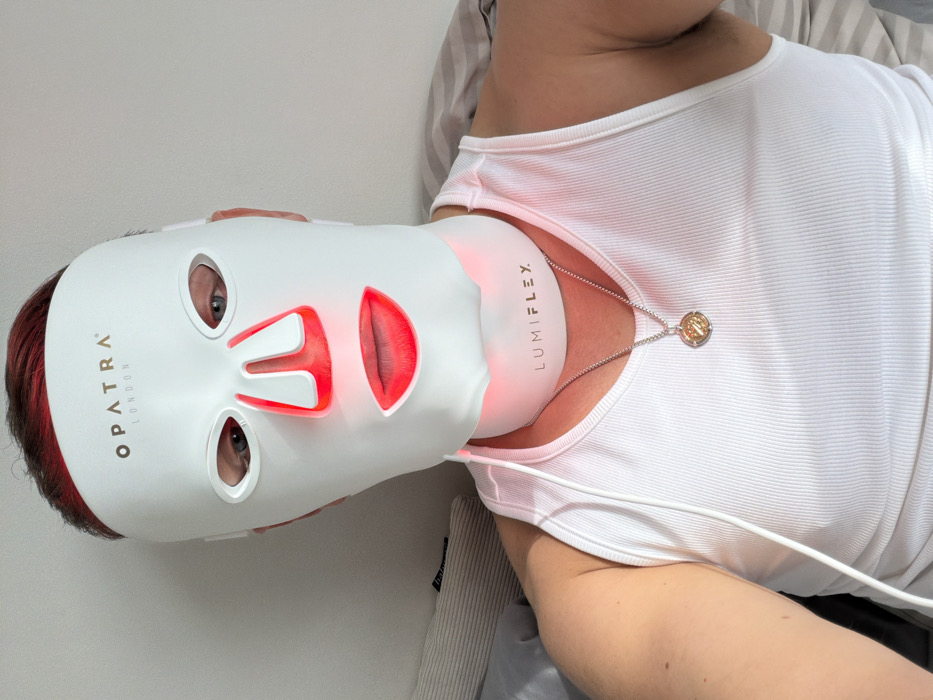 A person with short, reddish hair is depicted from the chest up in an indoor setting. They are wearing a white "OPATRA LONDON" branded LED facial mask, which emits red light around the nasal and oral areas. Immediately below, a white "LUMIFLEX" branded LED neck device is worn, also emitting red light. A white power cable extends from this neck device. The individual wears a white ribbed tank top and a silver-colored chain necklace with a circular pendant featuring a gold-colored emblem. The background consists of a plain, light-colored wall or surface, with a striped pillow or fabric visible on the left. The branding "LONDON" on the facial mask indicates an association with London, United Kingdom.