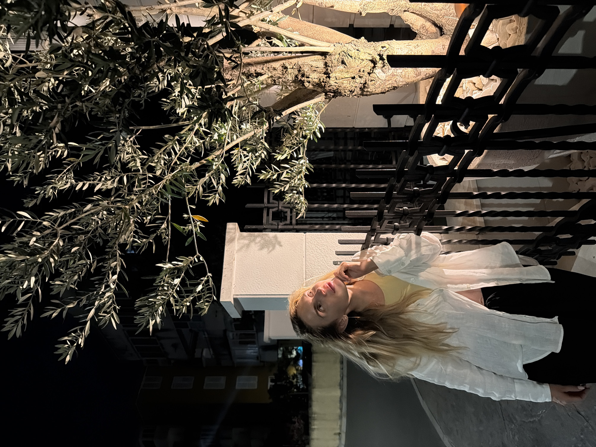 A medium shot shows a young blonde woman in a white open shirt and black pants standing outdoors at night. She is looking up and to the right, with her right hand touching her chin. A tree with illuminated branches is to her right, behind a black metal fence. In the background, there are buildings and a street. The scene appears to be in a courtyard or garden area in Budva, Montenegro.