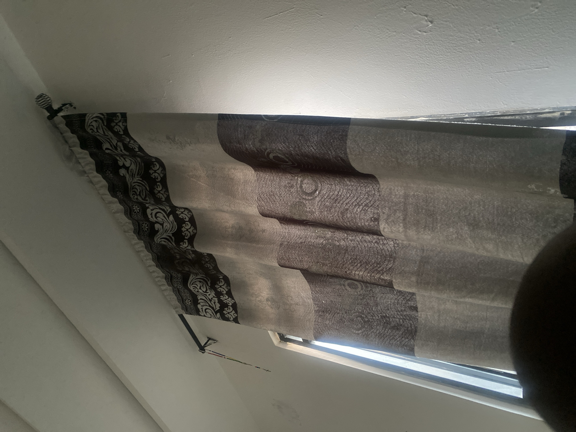 This image presents an upward-angled interior view of a room, specifically a window partially obscured by a curtain. The location is identified as Jalingo, Nigeria.
The central subject is a single curtain panel, exhibiting a multi-patterned design. The top section of the curtain features a wide band of dark fabric adorned with a complex white or light-grey scrollwork pattern, resembling acanthus leaves and fleurs-de-lis. Below this, the curtain fabric transitions to a lighter, semi-translucent material with subtle wavy and circular textures, followed by a lower section of darker fabric displaying prominent circular and wave-like patterns. The curtain is gathered into vertical folds and hangs from a black curtain rod.
The black curtain rod is mounted above the window. Its right end terminates in a spherical finial with a distinct ribbed or spiral texture. On the left side of the curtain rod, near its mounting bracket, a multi-colored beaded string, consistent with a rosary, is suspended. The beads are various colors, including red, green, yellow, and blue, culminating in a small crucifix or charm.
A portion of the window is visible on the left, behind the curtain. The window frame appears dark, and bright daylight is evident beyond the glass, indicating an outdoor light source. The surrounding walls and ceiling are painted a uniform white or off-white. These surfaces exhibit minor textural inconsistencies, potential smudging, and subtle wear or unevenness, particularly on the wall area to the left of the window. A discernible seam where the ceiling meets the wall is visible in the upper left.
At the bottom center of the frame, a dark, out-of-focus, curved mass is present, consistent with the upper part of a human head or shoulder in the immediate foreground. No individuals are clearly identifiable within the primary plane of focus.