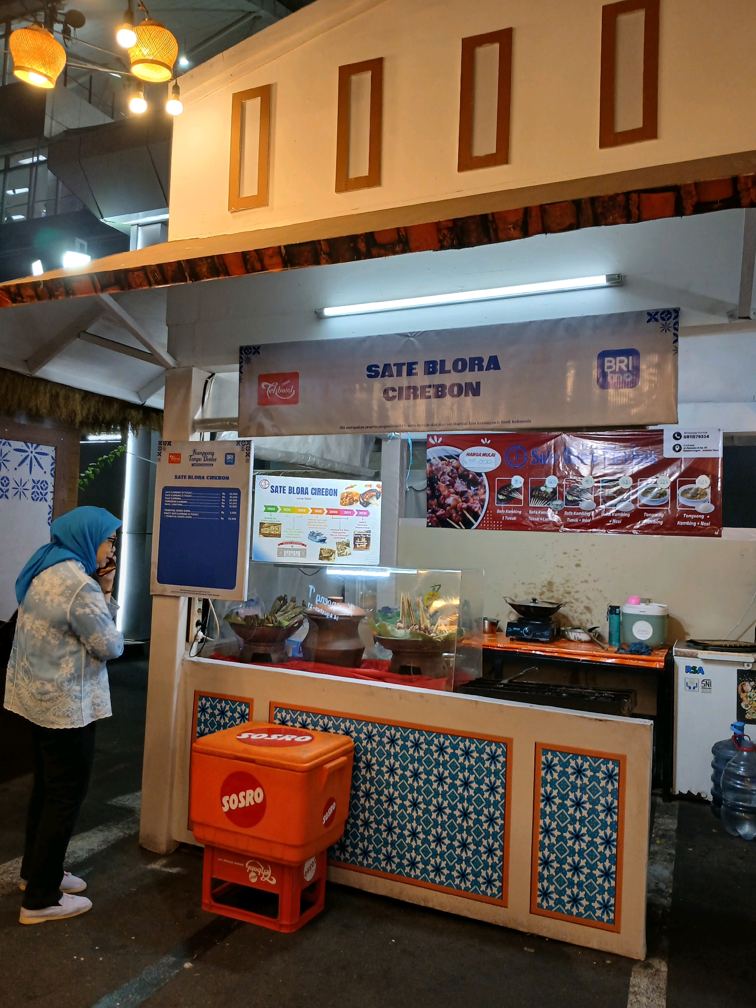 The image depicts an outdoor food stall in North Jakarta, Indonesia, specializing in "Sate Blora Cirebon." The stall is brightly lit by overhead bamboo-style lamps and a fluorescent tube light. A banner above the stall reads "SATE BLORA CIREBON" with a BRI (Bank Rakyat Indonesia) logo. A woman wearing a blue hijab and a patterned blouse is standing to the left of the stall, looking towards a menu board. The menu board displays prices for various sate dishes. To the right of the stall, there is a red cooler box with the "SOSRO" logo, placed on an orange plastic crate. The stall itself features a display counter with food items and cooking equipment, including pots and a stove. Behind the counter, a refrigerator with "RSA" and "SNI" labels is visible, along with large water cooler bottles. The stall's facade is decorated with a patterned tiling. The overall environment suggests a bustling evening market or street food setting.