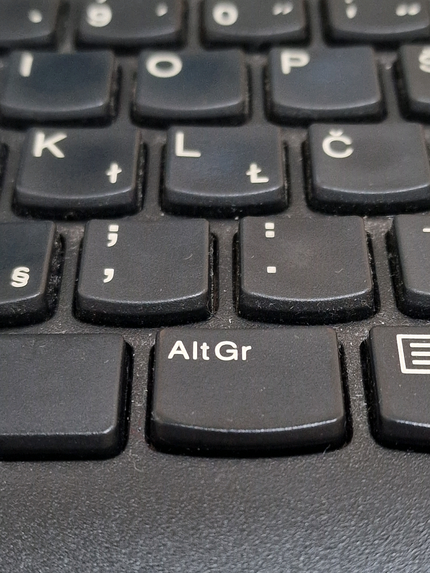 A close-up, overhead shot shows a black laptop keyboard. The image is focused on a few rows of keys, including the "Alt Gr" key which is clearly visible and labeled. Other visible keys include "K", "L", "P", "Č", ";", ",", and "§". The keys are slightly dusty, indicating the keyboard is in use. The overall color palette is dark, dominated by the black of the keys and the textured surface of the laptop. The background is dark and out of focus, suggesting a dimly lit environment. There are no people or any other objects visible in the frame.