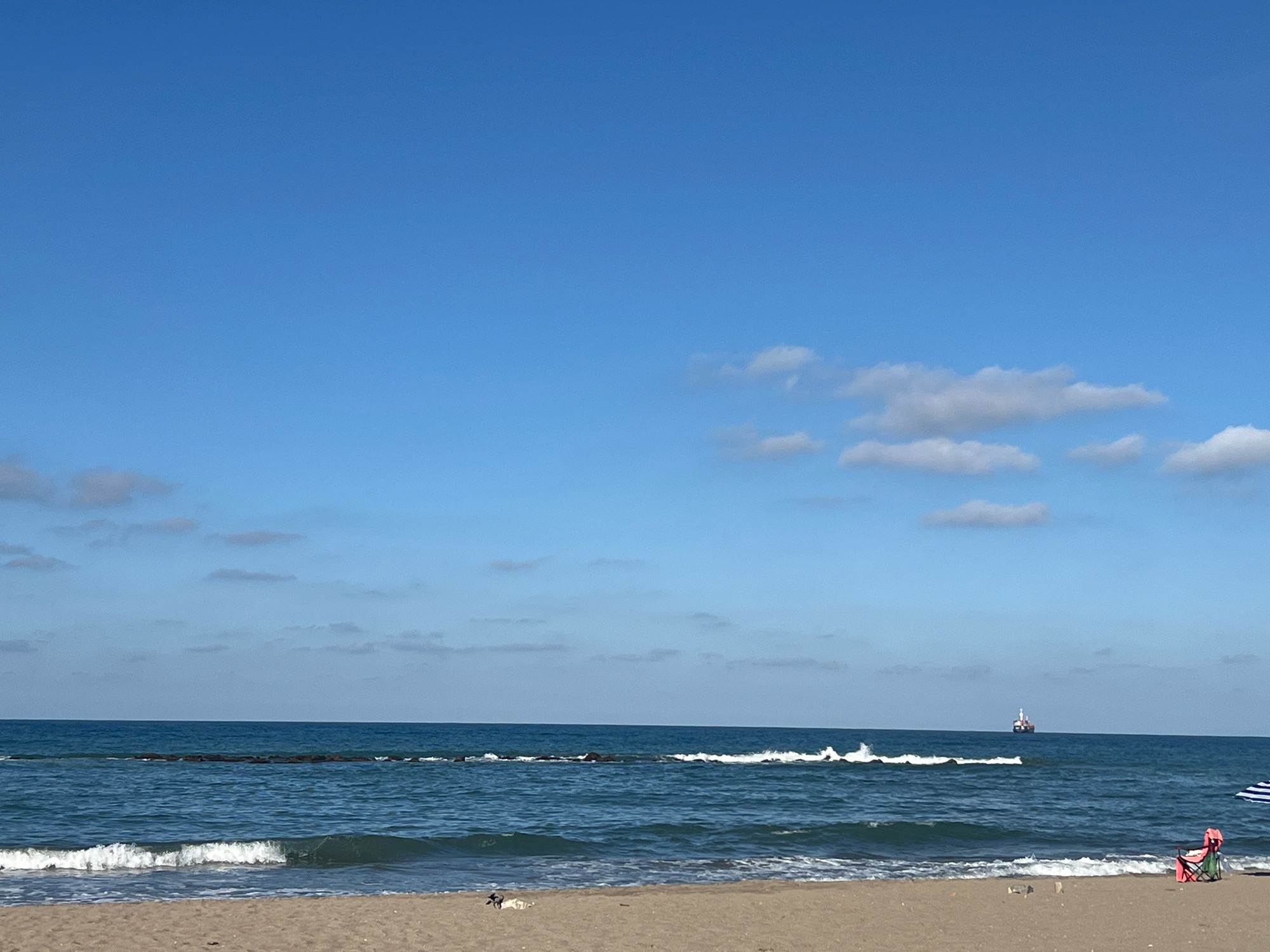 The image depicts a serene beach scene in Karasu, Turkey, under a clear blue sky dotted with scattered clouds. A calm sea stretches towards the horizon, where a distant cargo ship is visible. Gentle waves break along the shoreline, lapping onto a sandy beach. In the foreground, a person sits in a pink beach chair under a blue and white umbrella. The beach appears sparsely populated, with a few scattered objects and a bird near the water's edge. The overall atmosphere is one of tranquility and relaxation.