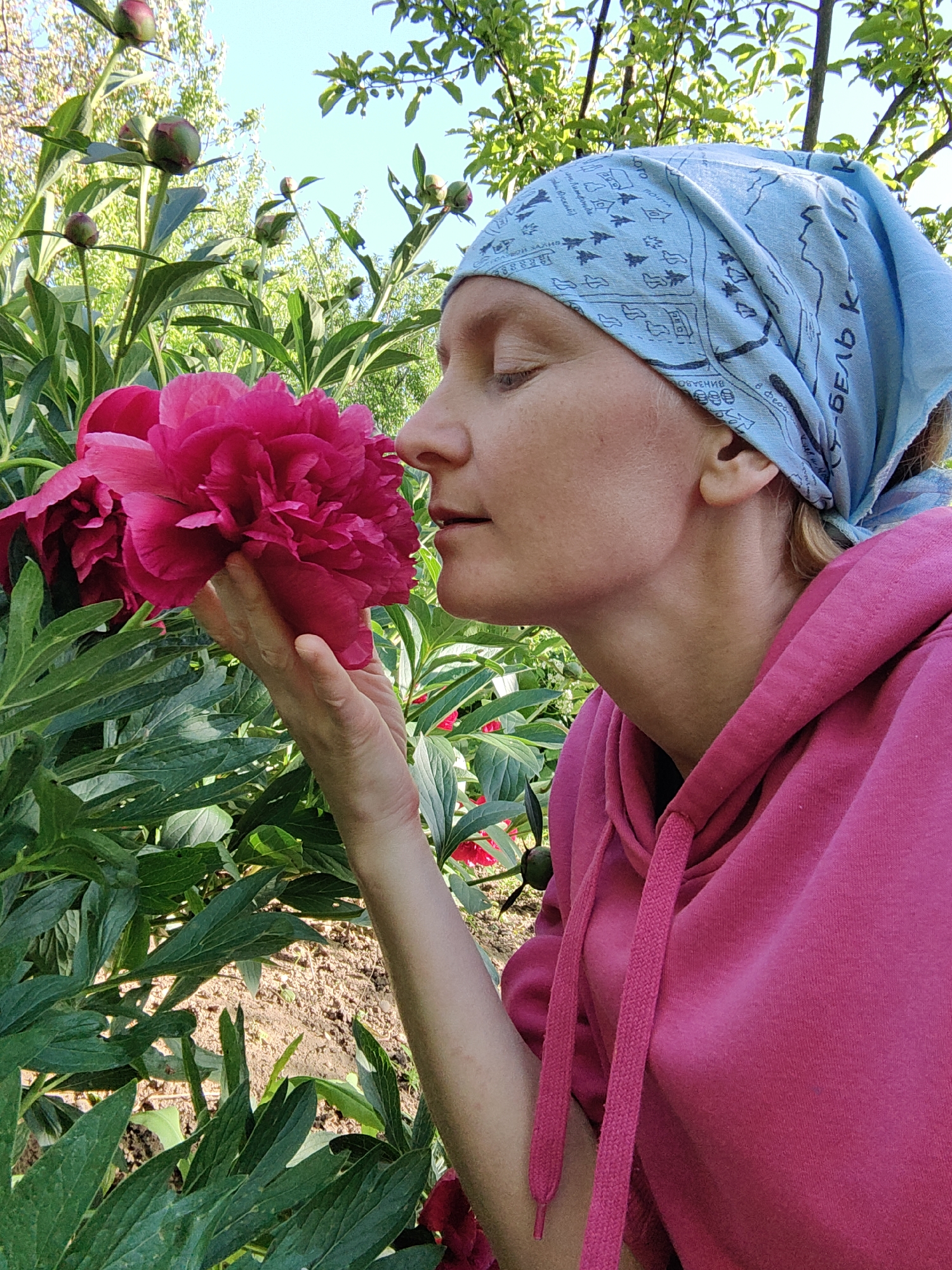 A fair-skinned woman with a light blue patterned bandana is smelling a vibrant pink peony flower in an outdoor garden setting. The woman is wearing a pink hooded sweatshirt. The peony is densely petaled. Green leafy plants surround the flower. The ground appears to be soil. Above the plants and woman, the sky is light blue and visible through the trees.