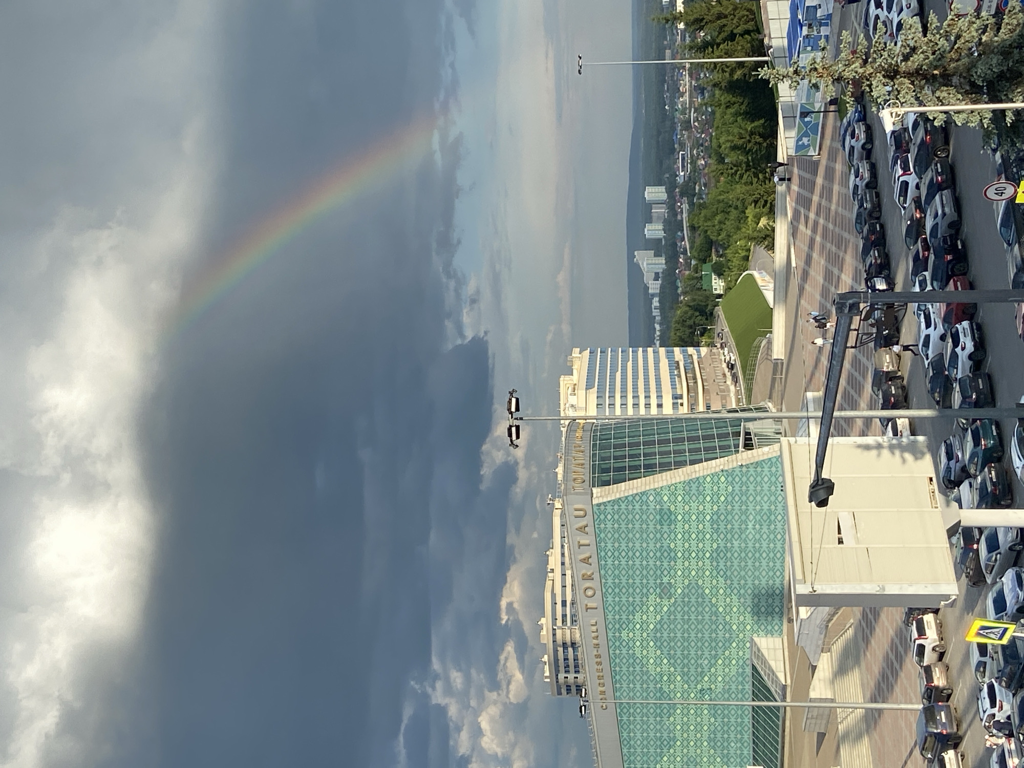 The image captures a wide-angle, high-angle view of the Congress-hall Toratau in Ufa, Russia. A vibrant rainbow arcs across a dramatic sky filled with dark storm clouds contrasted by patches of bright sunlight. In the foreground, the distinctive green patterned facade of the Congress-hall Toratau is visible, with the text "CONGRESS-HALL TORATAV" prominently displayed. Adjacent to the hall are modern white buildings. Below, a large parking lot is filled with numerous cars of various makes and models. A few individuals are seen walking in the plaza area in front of the hall. The background reveals a cityscape with more buildings and lush green trees, suggesting a suburban or park-like setting within the city. A lamppost stands on the right side of the frame, and a large pine tree is also visible. The overall scene suggests a post-rain event with a rainbow appearing after a storm.