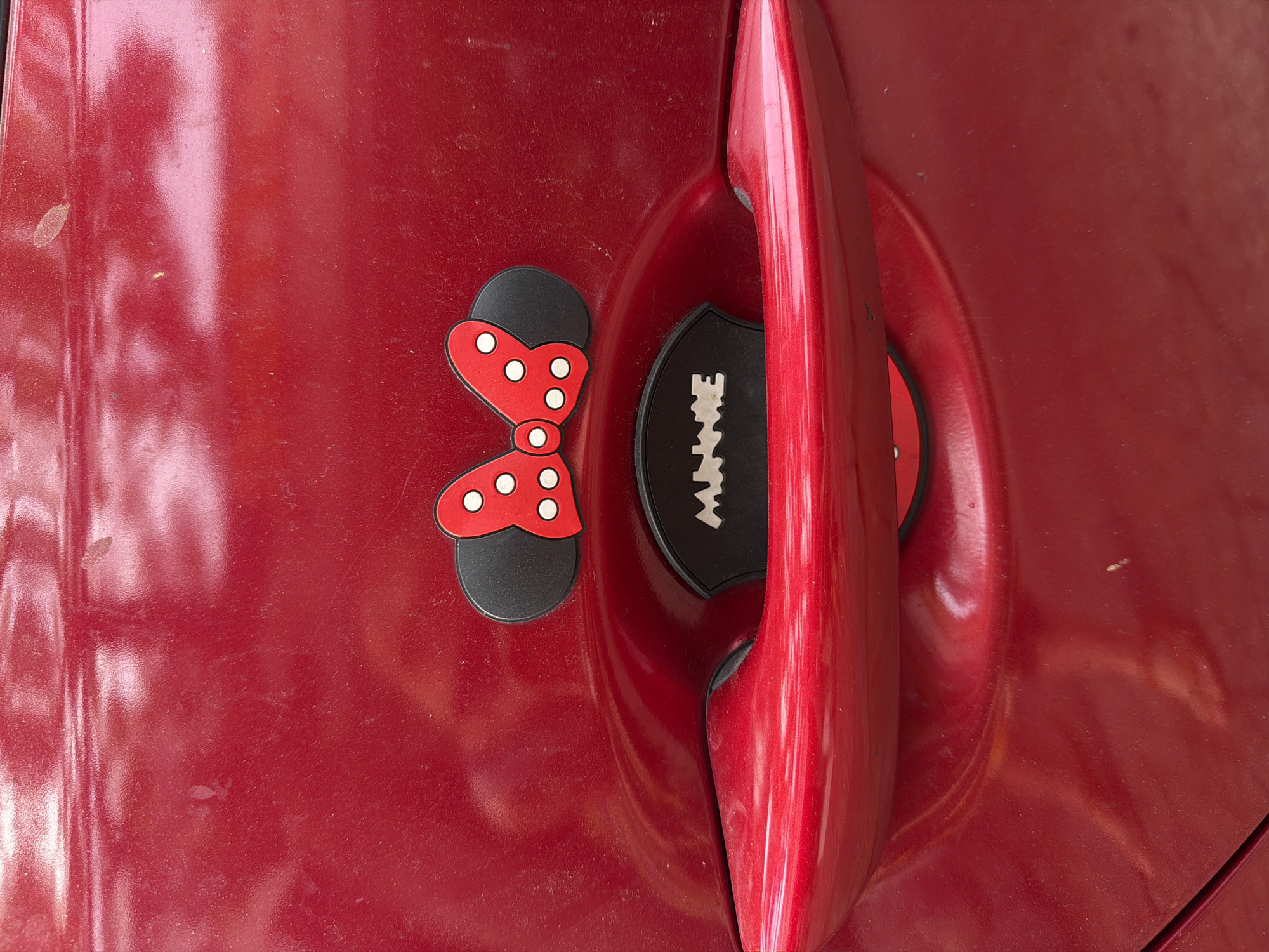 A close-up, medium shot captures a red car door with a chrome handle. Adorning the door are two decorative Minnie Mouse ears, each featuring a black ear with a red bow accent adorned with white polka dots. Below the ears, a black oval sticker is visible, emblazoned with the white letters "MINNIE". The car door shows signs of wear, with light scratches and dust visible on its surface. The scene is devoid of any people or surrounding environmental details, suggesting it was taken in an unknown location.