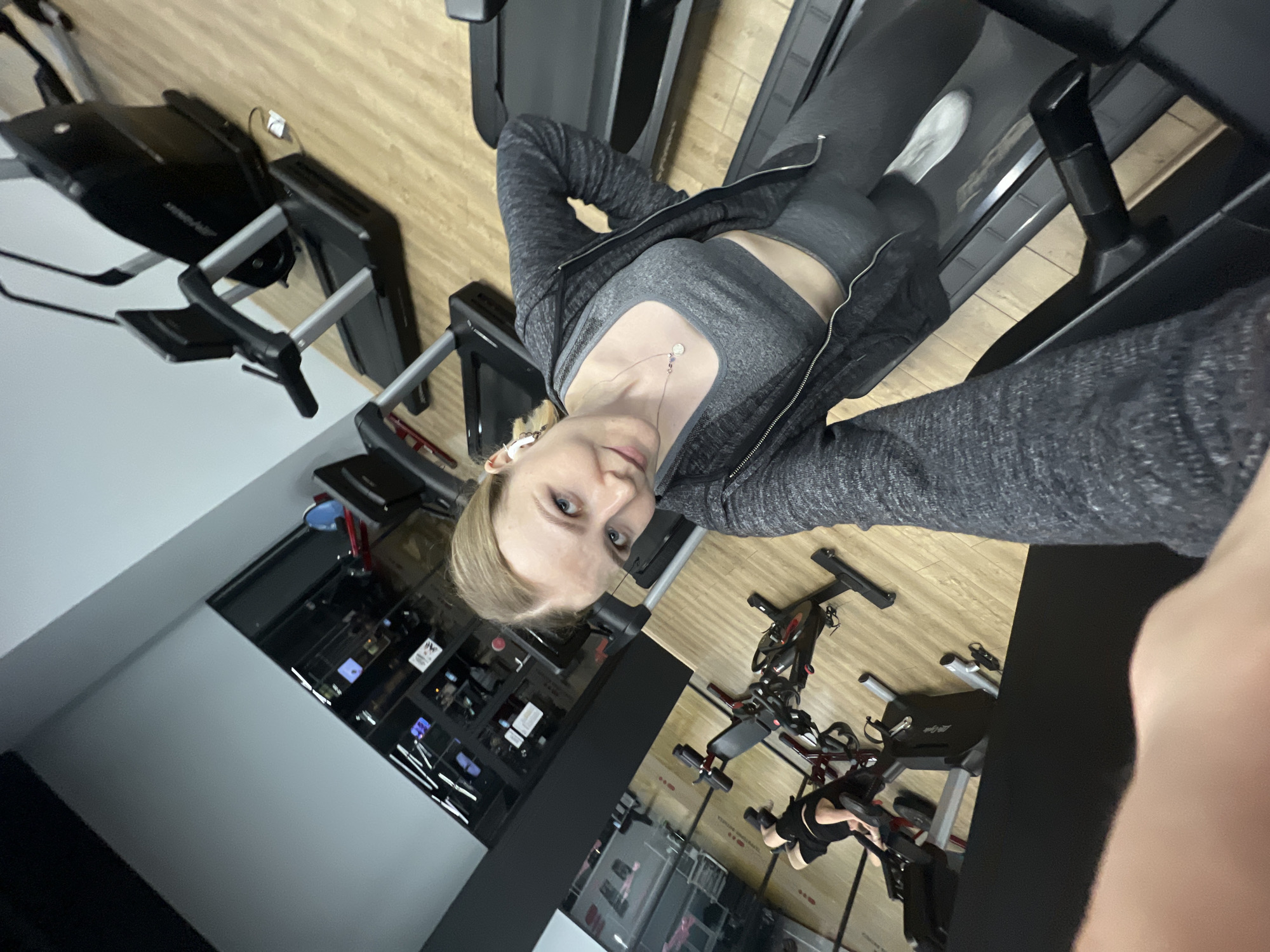 A fair-skinned woman in gray workout attire is taking a selfie while standing on a treadmill at a gym. The gym features treadmills, stationary bikes, and other fitness equipment. The location of the gym could be Khimki, Russia, because of a reflective sign that could contain the word "Khimki."