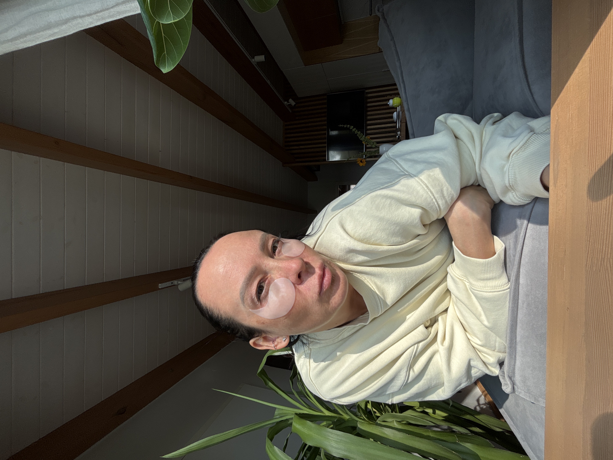 A medium, angled indoor shot depicts a person with eye patches lounging on a gray sofa. The individual is fair-skinned with dark hair pulled back, and wears a light yellow sweater. They are looking at the camera with a slight smile. Two pink eye patches are applied under their eyes.
The background shows a modern interior with wood-beamed ceilings and white paneled walls. A large green plant is visible in the lower portion of the frame. Deeper in the background, a television screen is visible. There is a wooden beam in the foreground.
The location and the details of the city Chaikovsky, Russia are not detectable in the image.