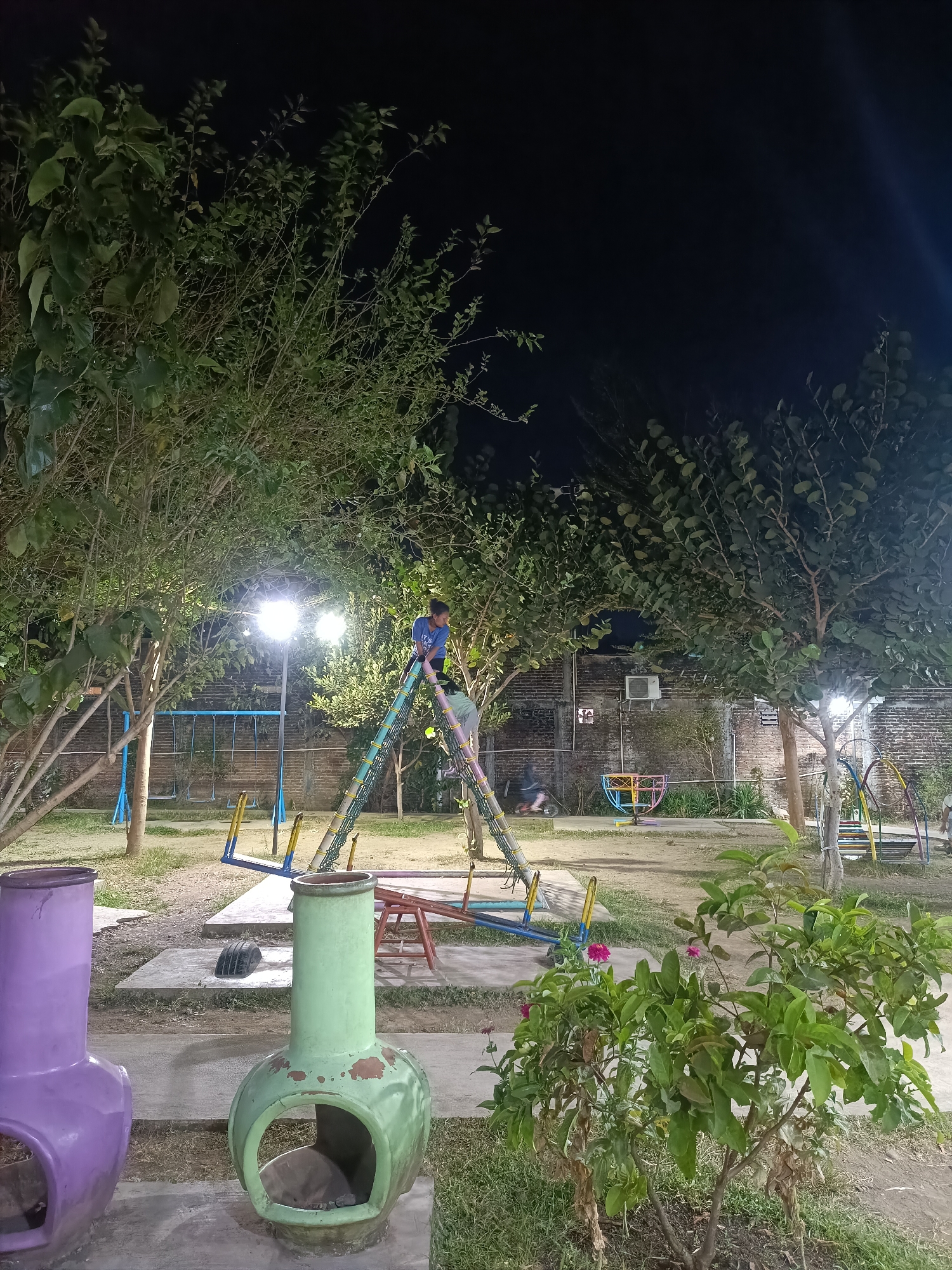 The image depicts a playground area at night in the city of Yogyakarta, Indonesia. A young boy in a blue shirt is climbing on a colorful A-frame structure. To the left, a set of swings with blue frames and yellow seats is visible, along with other playground equipment in the background. The ground is covered in dry grass and dirt. Two decorative outdoor fireplaces, one purple and one green, are in the foreground. Trees with dense foliage surround the playground, and a brick wall forms the background. Lighting from street lamps illuminates the scene.