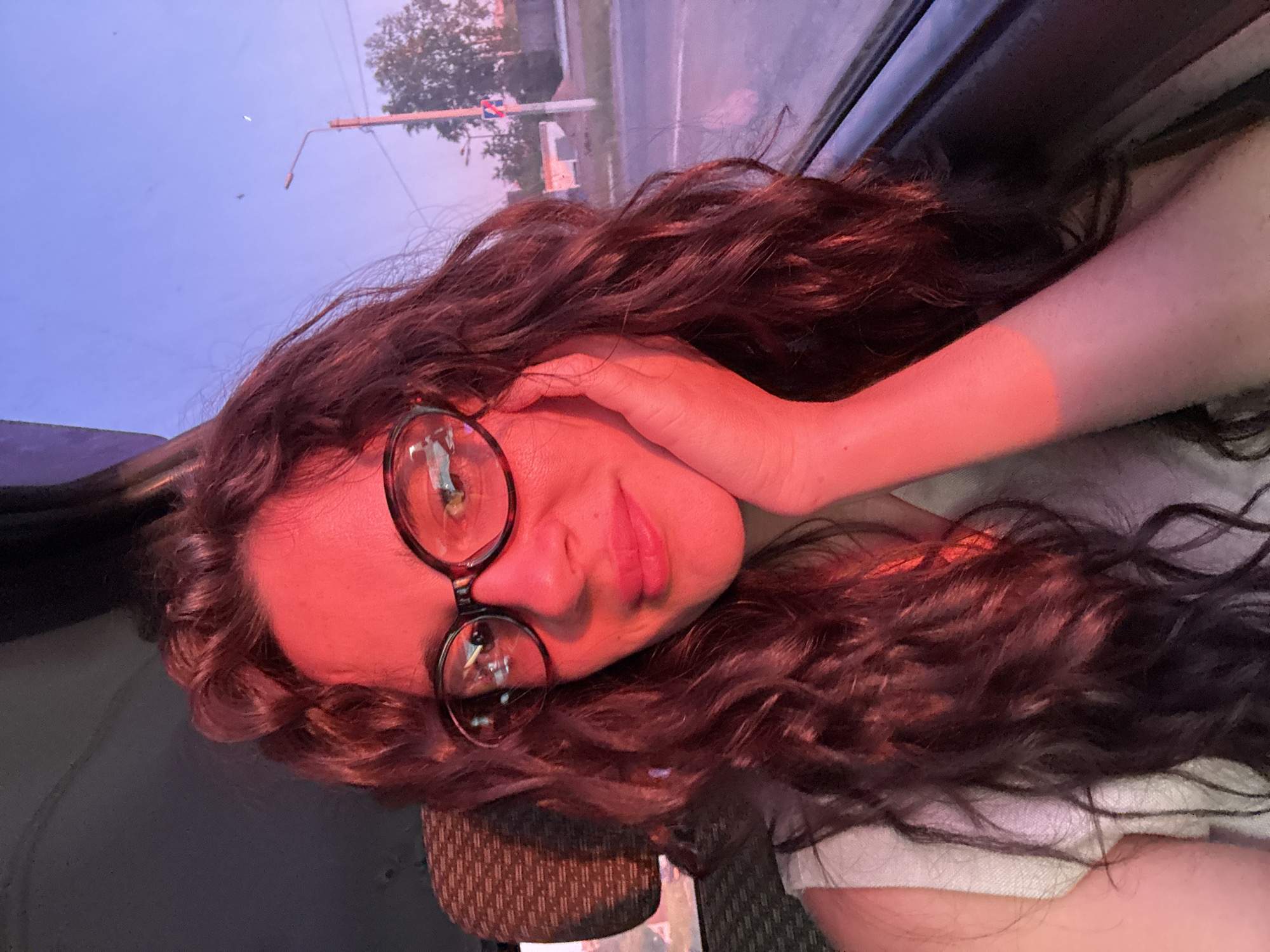 A woman with long, curly brown hair and eyeglasses is reclining, possibly in a vehicle, as suggested by the window and partial interior visible in the background. Her left hand is positioned near her face. The scene is illuminated by a reddish light, possibly from an external source.
Through the window, a street scene is visible with a traffic light, street lamp, and trees. Considering the streetscape, the location is plausibly Chaikovsky, Russia as the person is located in Chaikovsky, Russia.