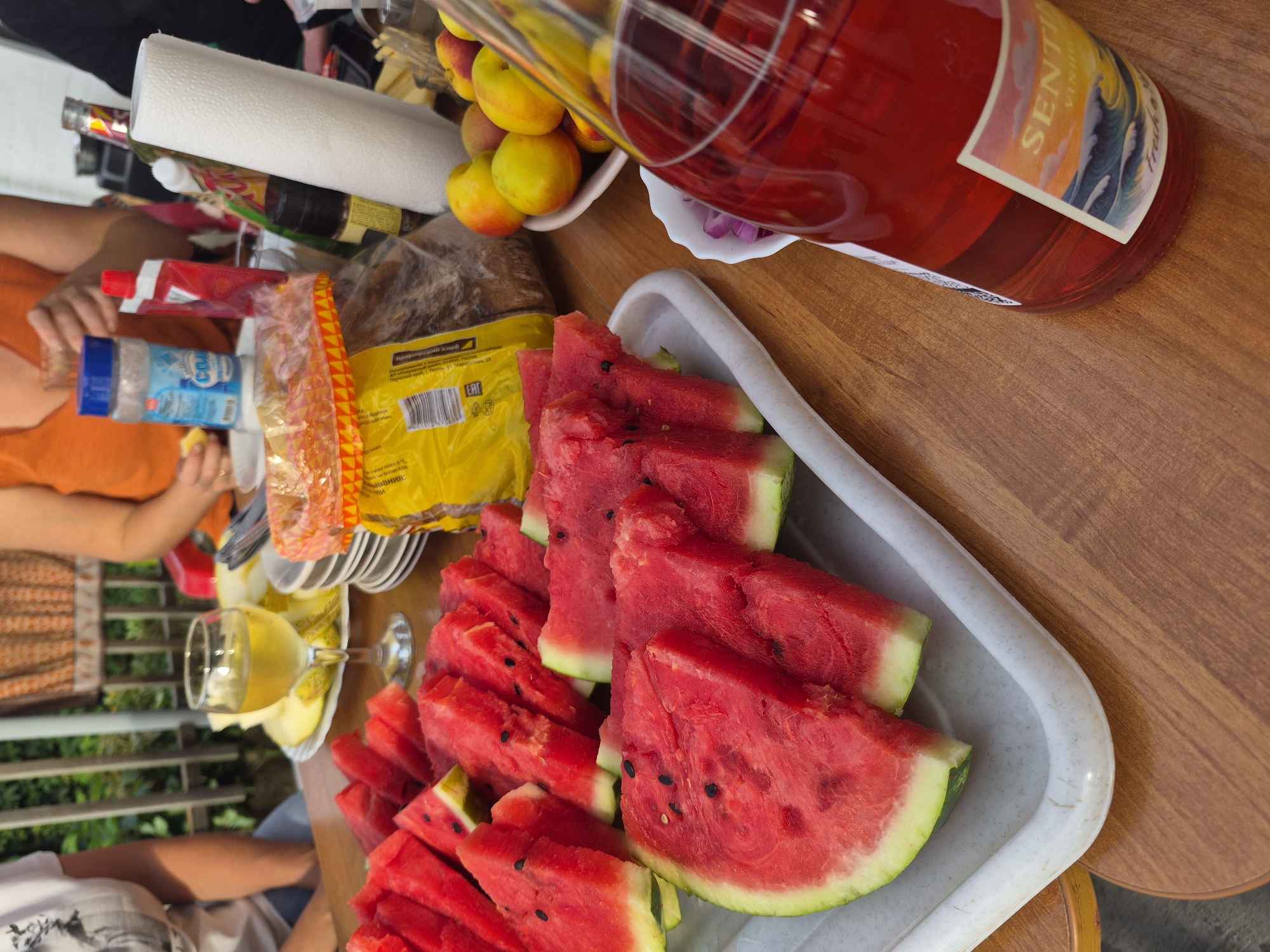 The image depicts an outdoor gathering, likely a picnic, set on a wooden table. A variety of food and drinks are laid out, including sliced watermelons on a white tray, a glass of white wine, a bottle of red liquid with a label that reads "SENTI VINHO," a bowl of nectarines, and a package of bread. In the background, blurred figures of people can be seen, one holding a glass and another wearing an orange top. The setting appears to be a backyard or park, with greenery and a wooden fence visible in the distance. The overall atmosphere suggests a casual and relaxed social event. The photo was taken in Chaikovsky, Russia.