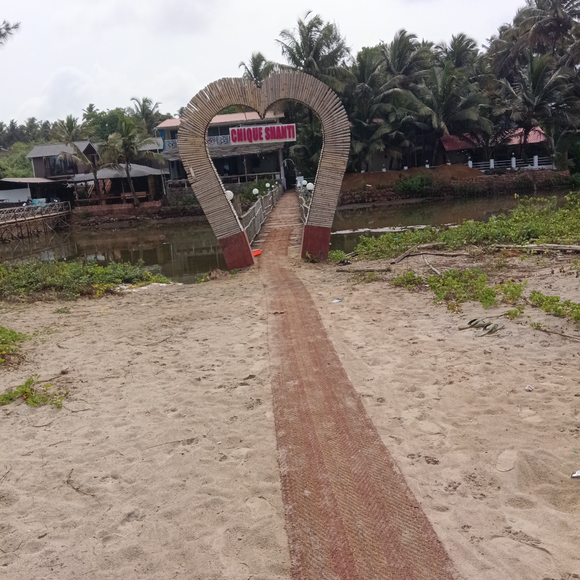 The image depicts a landscape in Mandrem, India, featuring a beachfront leading to a small bridge that crosses a body of water. Above the bridge is a heart-shaped bamboo archway, adorned with the sign "Chique Shanti."
The scene includes a sandy beach, a path made of what appears to be a woven material leading to the bridge, and calm water separating the beach from the building and trees beyond. The architecture indicates a tropical or coastal design. The presence of coconut trees contributes to the tropical ambiance. The sky is overcast, suggesting a cloudy day.