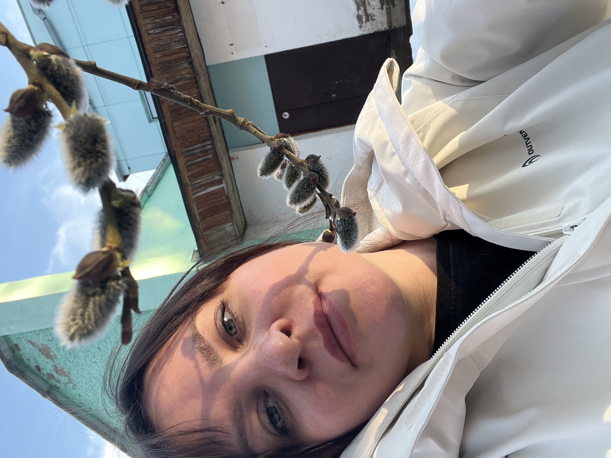 A woman takes a selfie outdoors, holding a branch of pussy willow. She is wearing a light-colored jacket. The background includes a light blue building with a brown door, indicating an urban environment, potentially Noviy, Russia.