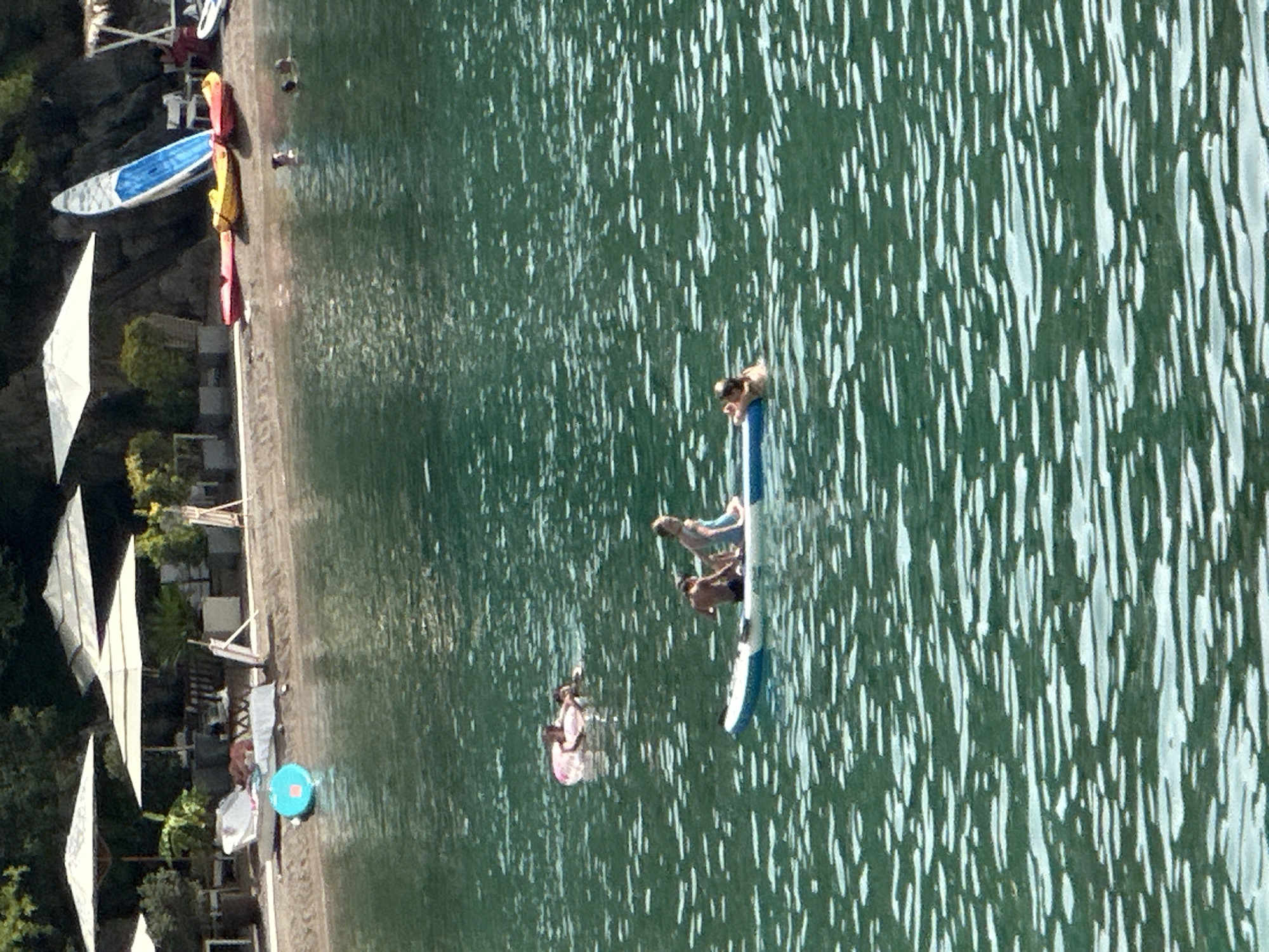 The image depicts a serene coastal scene, likely in Budva, Montenegro, as suggested by the context. In the foreground, a body of shimmering turquoise water is visible, its surface rippling with sunlight. Several individuals are engaged in water activities. A blue and white paddleboard is in the center, carrying three people: a man and two children. One child is sitting, while the other appears to be standing. To the left of the paddleboard, a pink inflatable ring floats on the water, with two people partially submerged and resting on it.
In the background, the shoreline is lined with sand and beach umbrellas. Various beach furniture, including lounge chairs and tables, are visible. A blue and white surfboard stands upright on the sand, leaning against the rocky embankment. Red and yellow kayaks are also resting on the shore. The scene is framed by lush green trees and vegetation on a hillside. The overall atmosphere is one of relaxation and recreation on a sunny day.