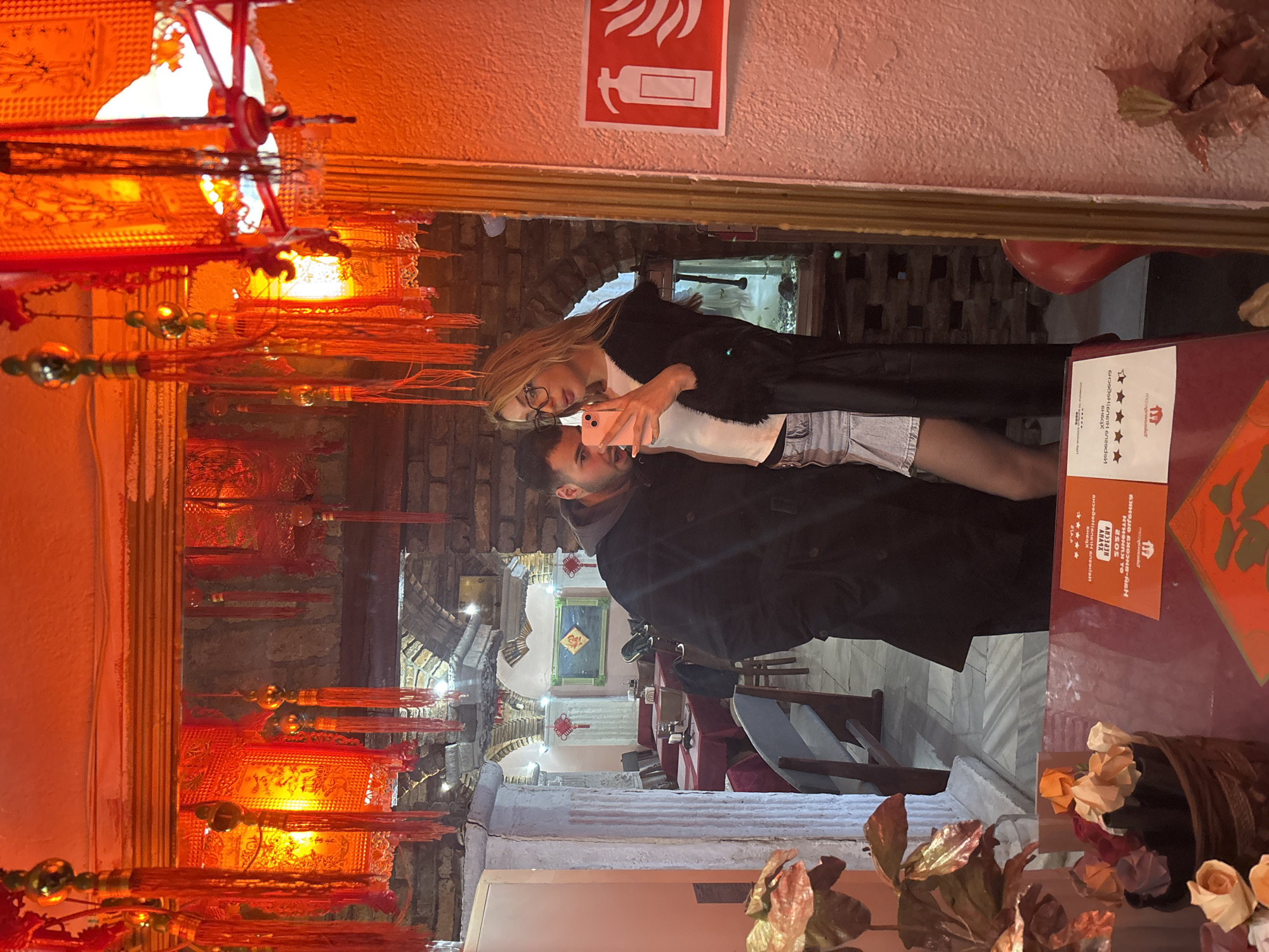 A couple is reflected in a mirror. The woman is fair-skinned with long blonde hair and wearing glasses, a white top, denim shorts, and a black fur-trimmed jacket. She holds a pink phone. The man has dark hair and a beard and wears a dark coat. The background behind the mirror suggests an Asian-themed restaurant or establishment with ornate red lanterns and decorations. A fire extinguisher sign is visible on the wall above the mirror. The restaurant setting, along with the rating card on the table, suggest the location is "Happy Sushi Varna" in Varna, Bulgaria.