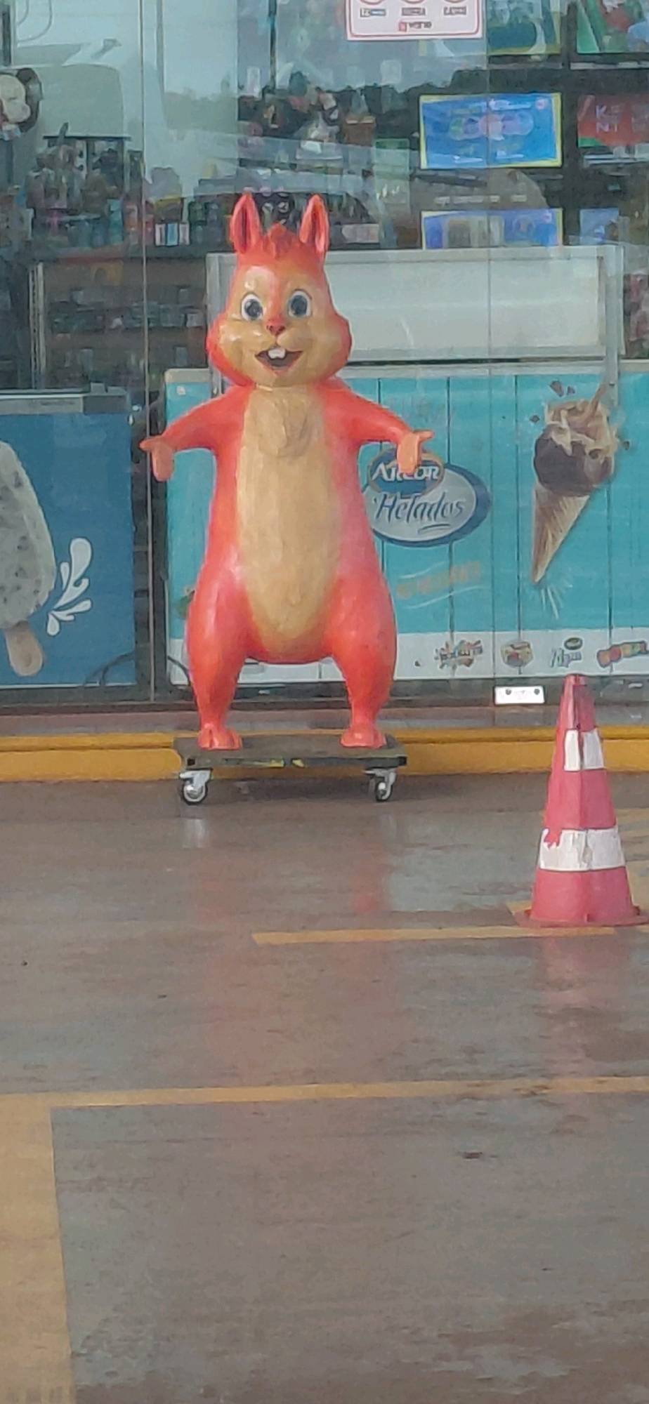 In Encarnación, Paraguay, a brightly colored, anthropomorphic squirrel statue is positioned outside an establishment. The statue, predominantly orange and yellow, stands on a wheeled platform, giving the appearance of riding a skateboard. It has large, expressive eyes, an open mouth with visible teeth, and prominent ears. Behind the statue, a blue wall features graphics of ice cream cones and the text "Alcor Helados," indicating it is likely an ice cream shop. A pink and white striped traffic cone is visible to the right of the statue. The ground appears to be paved with yellow lines marking lanes or parking spaces. The overall scene suggests an outdoor retail or recreational environment.