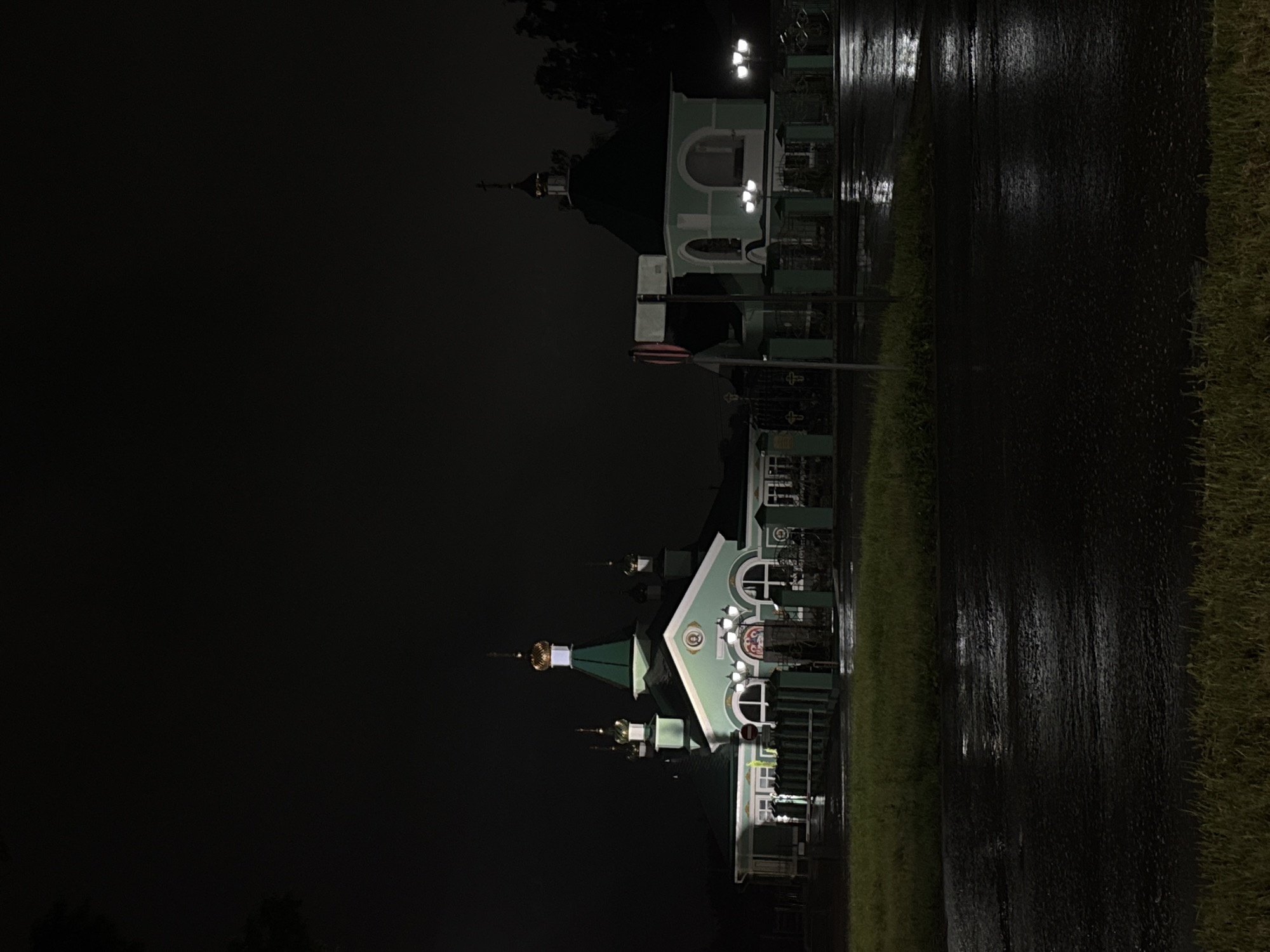 The image displays a religious architectural complex at night, located in the city of Chaikovsky, Russia. The scene is illuminated by artificial lighting, which reflects prominently on a wet, dark asphalt road in the foreground, indicating recent rain. The central structure is a building with a light green and white exterior, topped by dark green roofs and several golden-colored onion domes, most of which are surmounted by Orthodox crosses. The largest dome is centrally positioned over the main body of the building. The facade features arched windows and what appears to be a religious icon or mural above the primary entrance. The complex is enclosed by a dark fence with decorative elements and crosses on its posts. To the right of the main building stands a smaller, detached structure with a similar green roof and an open, arched entrance. A traffic sign, a red circle with a horizontal white bar, is partially visible in front of this secondary building. Multiple streetlights with white illumination are present, casting light onto the buildings and the reflective road surface. Patches of green grass are visible in the immediate foreground and between the road and the fenced property. The background is dark and indistinct, with tree silhouettes visible on the far right. No people are present in the image.