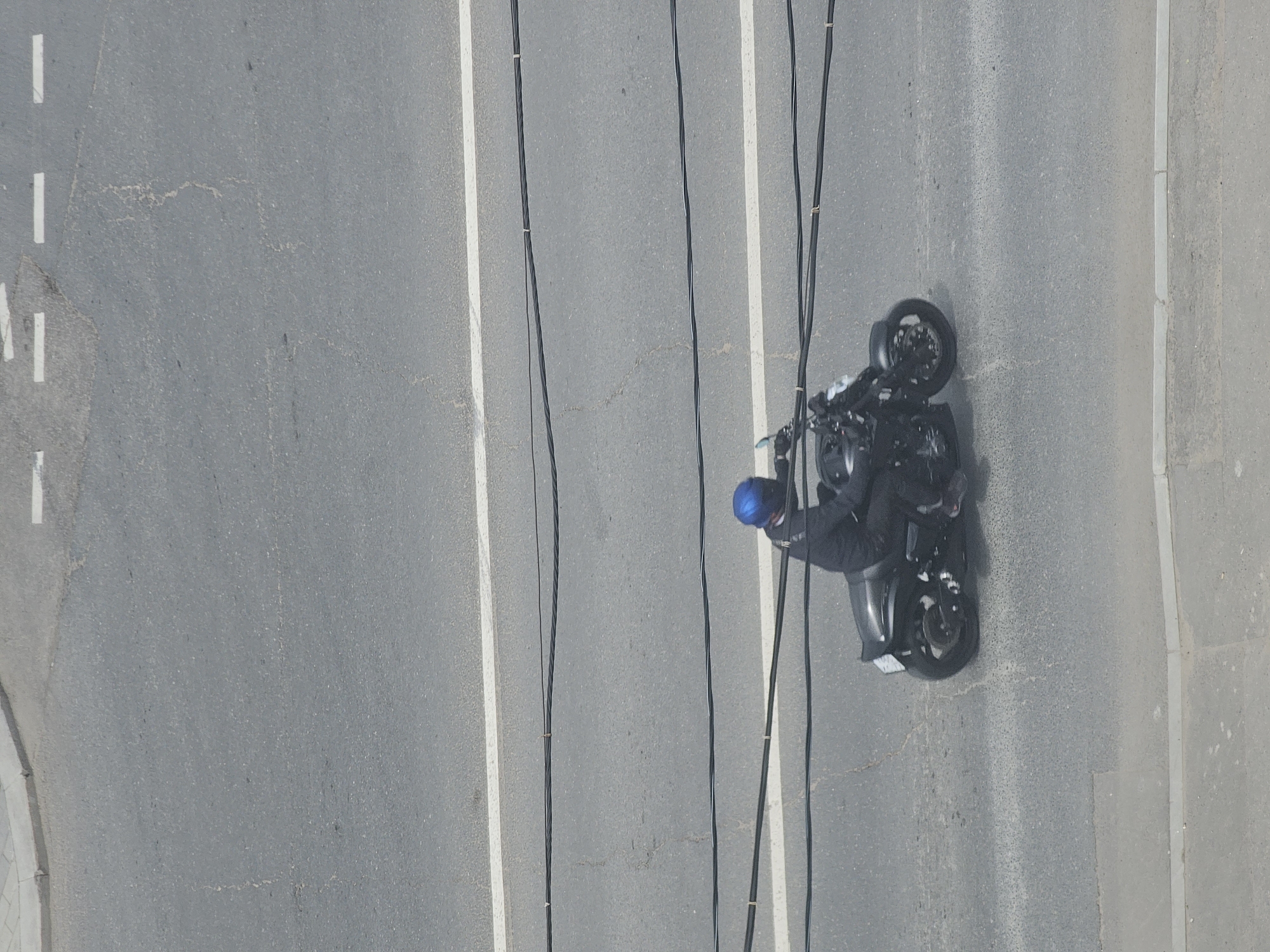 The image shows an aerial view of a motorcyclist on a road in Moscow, Russia. The motorcyclist is wearing a blue helmet and dark clothing, and is riding a black motorcycle. The road has white lane markings and is lined with trees. There are also power lines running across the scene. The lighting suggests it is daytime, and the weather appears to be clear. The overall scene is one of urban transportation. There is no visible text in the image.