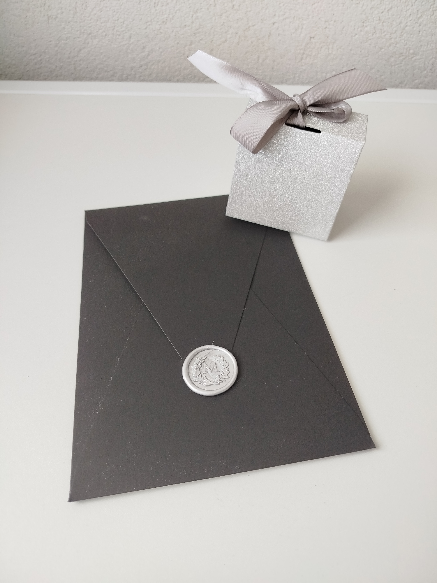 The image depicts a dark gray envelope with a silver wax seal bearing the letter "M," placed on a white surface. Next to the envelope is a small, sparkly silver box tied with a silver ribbon in a bow. The background is a plain, off-white wall. The objects appear to be on a flat surface, likely a table. There are no people visible. The location in the city of Varna, Bulgaria cannot be determined from the provided image.