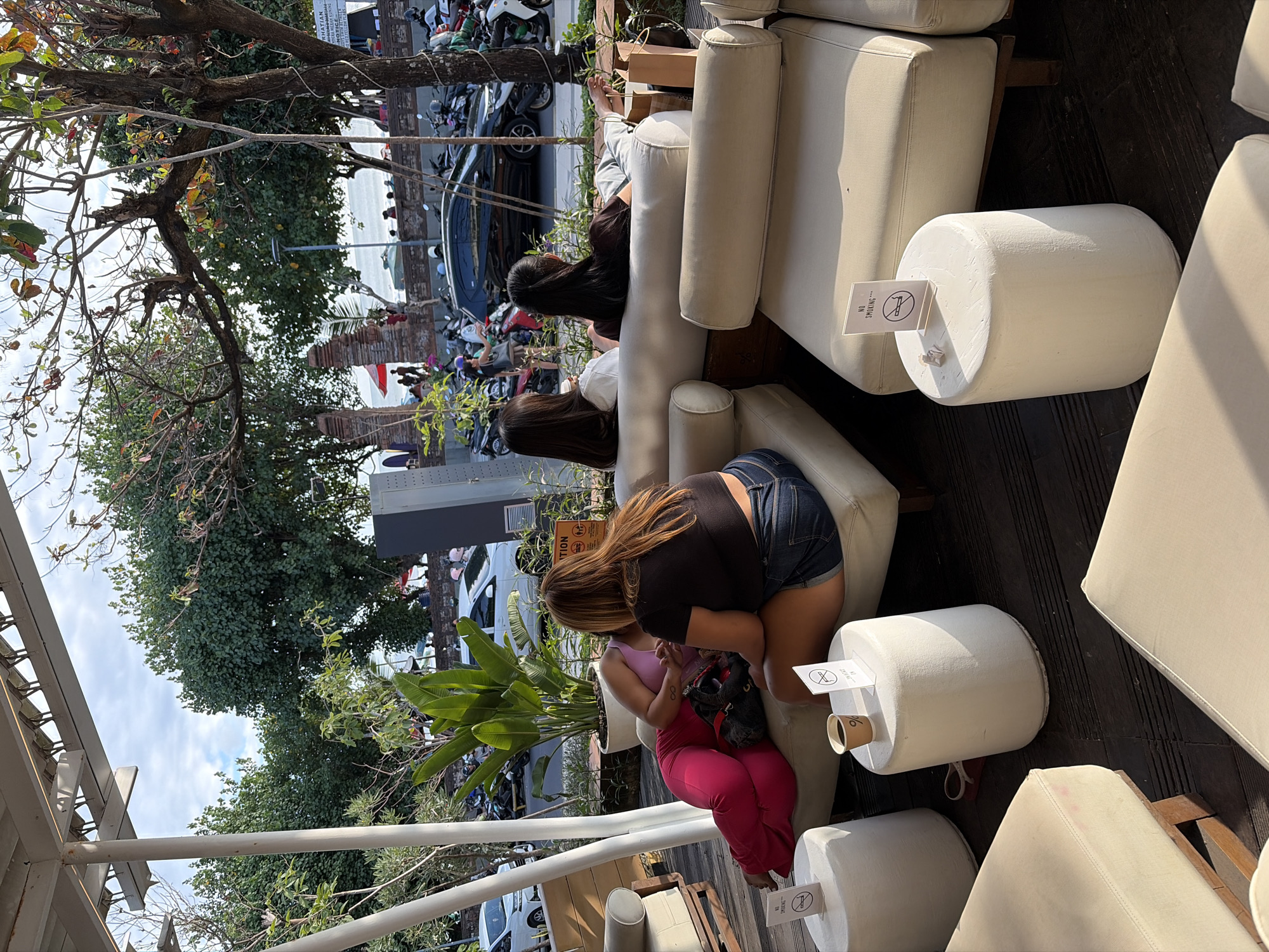 The image depicts an outdoor seating area, likely a cafe or restaurant, in Kuta, Indonesia. The setting features light-colored, modular sofas arranged on a dark wooden deck. Several round, white cylindrical stools are placed between the sofas. On one of these stools, a disposable coffee cup sits next to a small sign with a "no smoking" symbol.
In the foreground, three women are seated on the sofas. One woman, wearing bright pink pants and a purple top, is partially reclined. Another woman, in a black crop top and denim shorts, is seated facing away from the camera, looking towards the other two women. The third woman, also facing away, has long dark hair and is wearing a white top.
In the background, there is a street scene with parked motorcycles and cars, indicating an urban environment. Palm trees and other greenery are visible, suggesting a tropical location. Beyond the street, a beach and the ocean can be seen under a partly cloudy sky. People are visible on the beach and near a wall separating the seating area from the street. A prominent architectural feature, possibly a brick wall with decorative elements, is present in the mid-ground. The overall atmosphere suggests a relaxed, casual environment, common in tourist areas of Kuta.