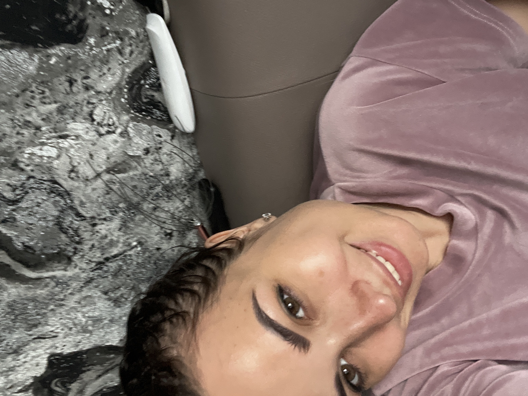 This media file is a close-up, slightly rotated, selfie of a young woman. She has dark hair, brown eyes, and is wearing makeup, including winged eyeliner. She is smiling, showing her teeth, and appears to be in a relaxed and happy mood. She is wearing a soft, light purple velour top. The background is partially visible and appears to be indoors, with a textured gray surface on the left and a brown surface on the right, possibly a sofa or chair. A white object, which resembles a remote control, is visible on the gray surface. The lighting suggests it could be daytime indoors.