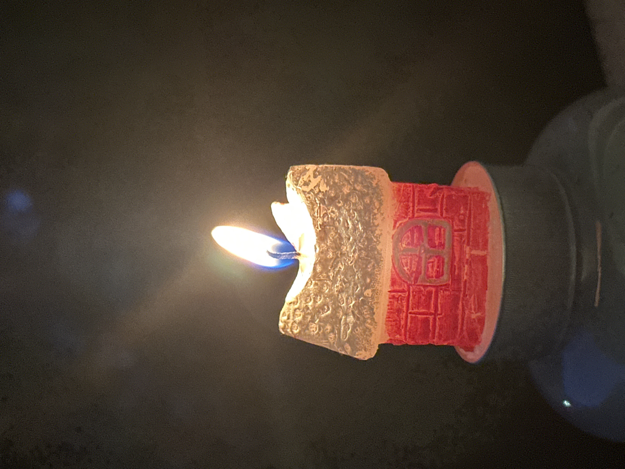 The image shows a close-up of a lit candle shaped like a small red house with a snow-covered roof. The candle is sitting on a dark blue base. The flame is bright and casts a warm glow, illuminating the intricate details of the house-shaped candle, including the brick pattern on the walls and a window. The background is dark and out of focus, creating a sense of intimacy and warmth. There is no indication of a specific location in Krefeld, Germany, or any people or interactions.