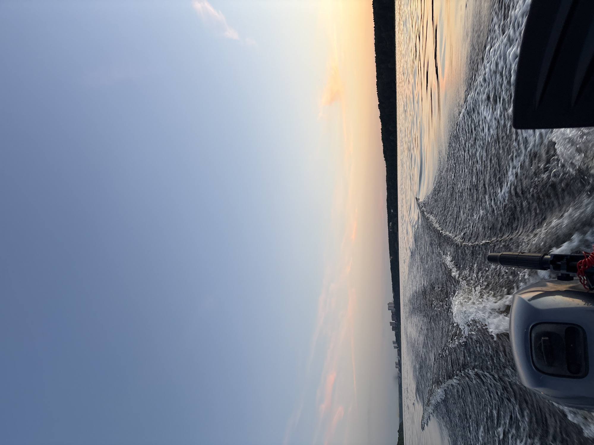 The image depicts a view from the back of a motorboat moving across a body of water, likely a river or lake. The water behind the boat is disturbed, creating a wake with white foam. In the foreground, the back of the boat's motor is visible, painted gray, with a red cord attached. Further in the distance, a line of trees forms the horizon, and beyond that, a city skyline with several tall buildings is visible. The sky is a gradient of light blue at the top, transitioning to pale pink and orange hues near the horizon, suggesting either sunrise or sunset. The overall impression is one of a peaceful journey on the water. The presence of a city skyline with multiple tall buildings in the distance suggests this could be a scene on the Kama River in Perm, Russia, given the context.
