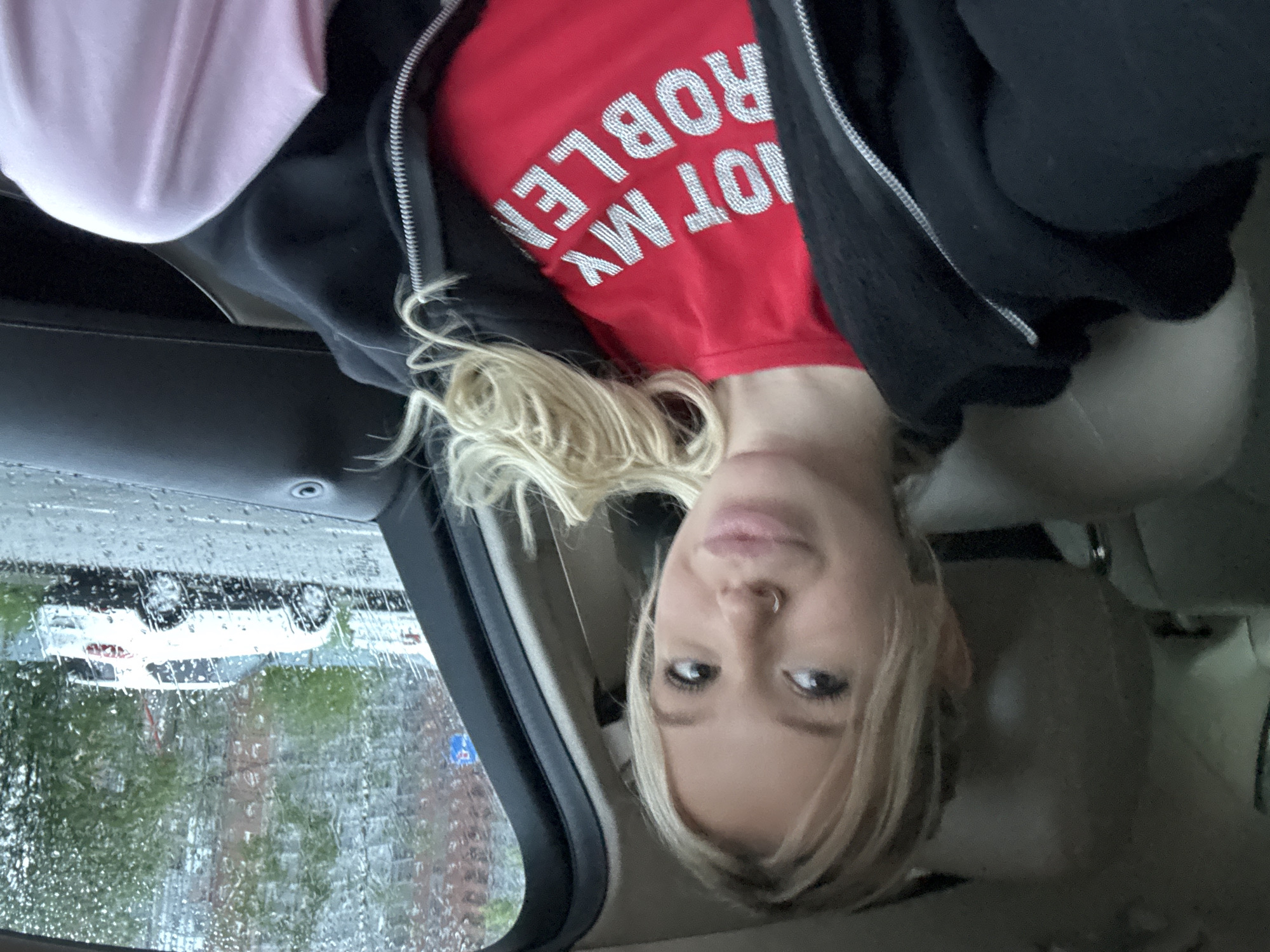 A young woman with blonde hair and a nose ring is sitting in the passenger seat of a car, looking towards the camera. She is wearing a red t-shirt with the words "NOT MY PROBLEM" written in rhinestones, and a black zip-up hoodie. The car is parked on a street in Saint Petersburg, Russia. It is raining, and the raindrops are visible on the car window. In the background, there are apartment buildings and trees. A white SUV is parked on the street.