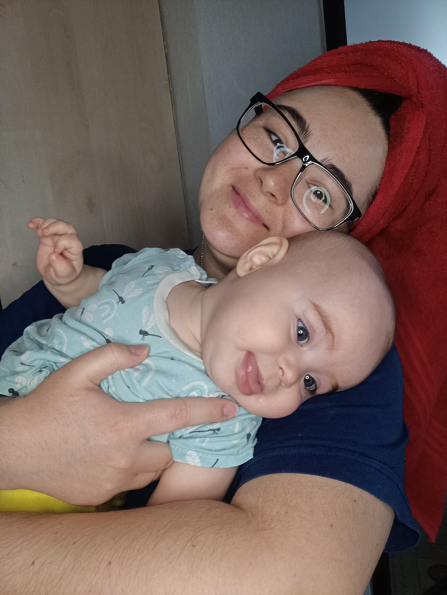 A selfie depicts a woman holding a baby. The woman wears black-framed glasses and has a red towel wrapped around her head. The baby is wearing a light blue outfit with a dragonfly pattern and is sticking its tongue out. The woman is holding the baby in her arms. The background is out of focus, but appears to be an interior setting. The picture was taken in Noviy, Russia.