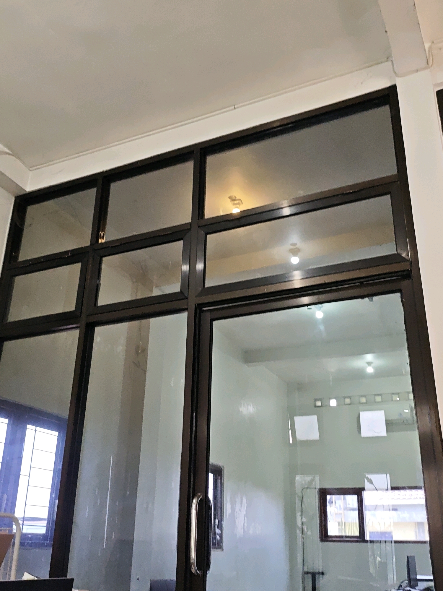 The image shows an interior view of a room with large glass windows and doors. The windows are divided into multiple panes by dark frames. Reflections on the glass show lights and possibly other rooms or buildings. A metal door handle is visible on one of the glass doors. The walls appear to be painted a light color. The overall scene suggests an indoor environment, possibly an office or a commercial space. The location provided, Ngasem, Indonesia, indicates the city where this scene is situated.