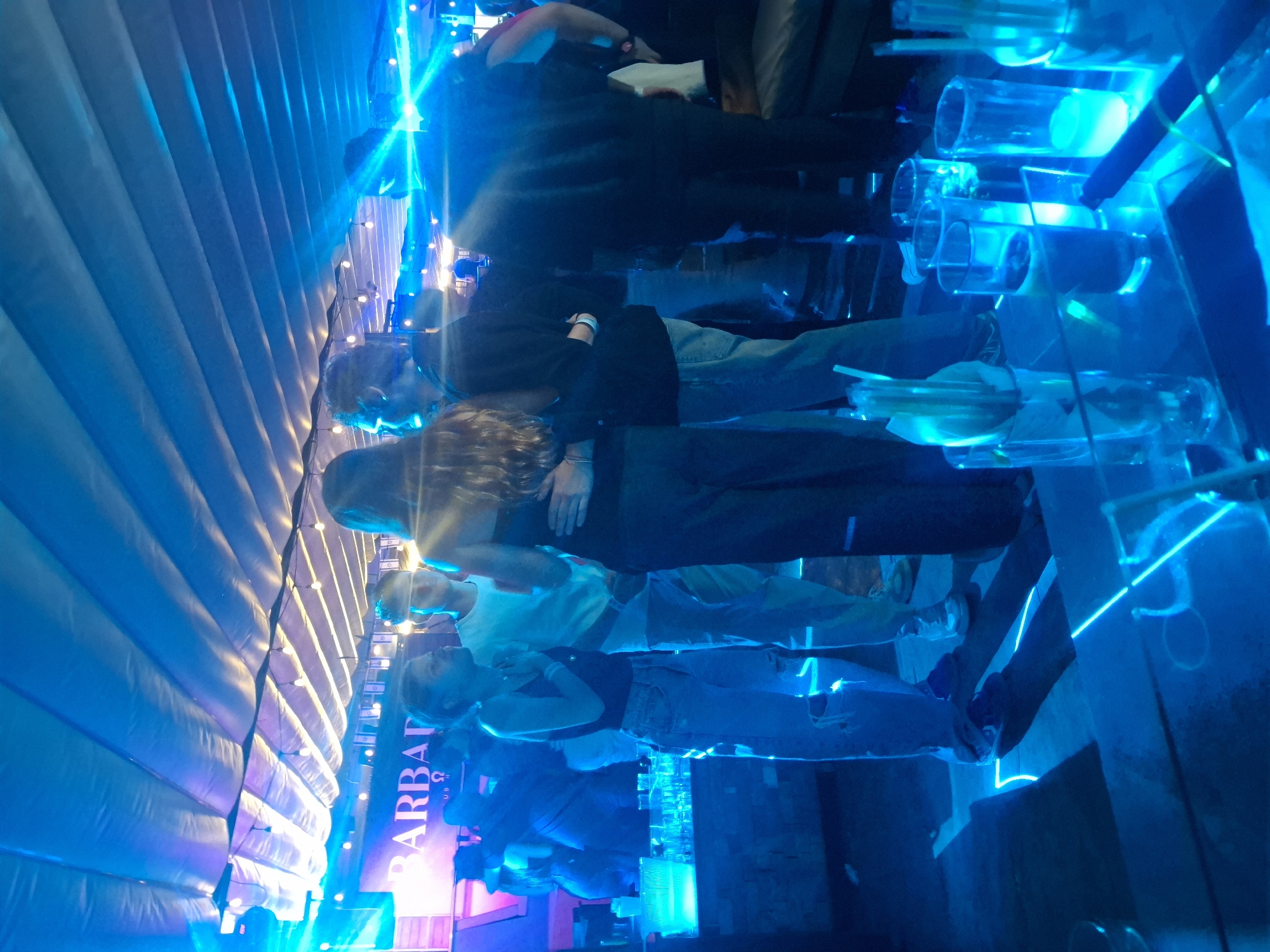 The image depicts a crowd of people, likely at a nightclub or bar in Mexico City, Mexico. The location is indicated by the word "BARBARA" appearing prominently in the frame. The environment is dominated by blue lighting. A display of illuminated, transparent cylinders containing objects, perhaps drinking glasses or bottles, is visible. People are standing and appear to be interacting with each other. Individuals are dressed in casual attire. The scene has an overall vibrant, energetic atmosphere, consistent with a nightlife setting.