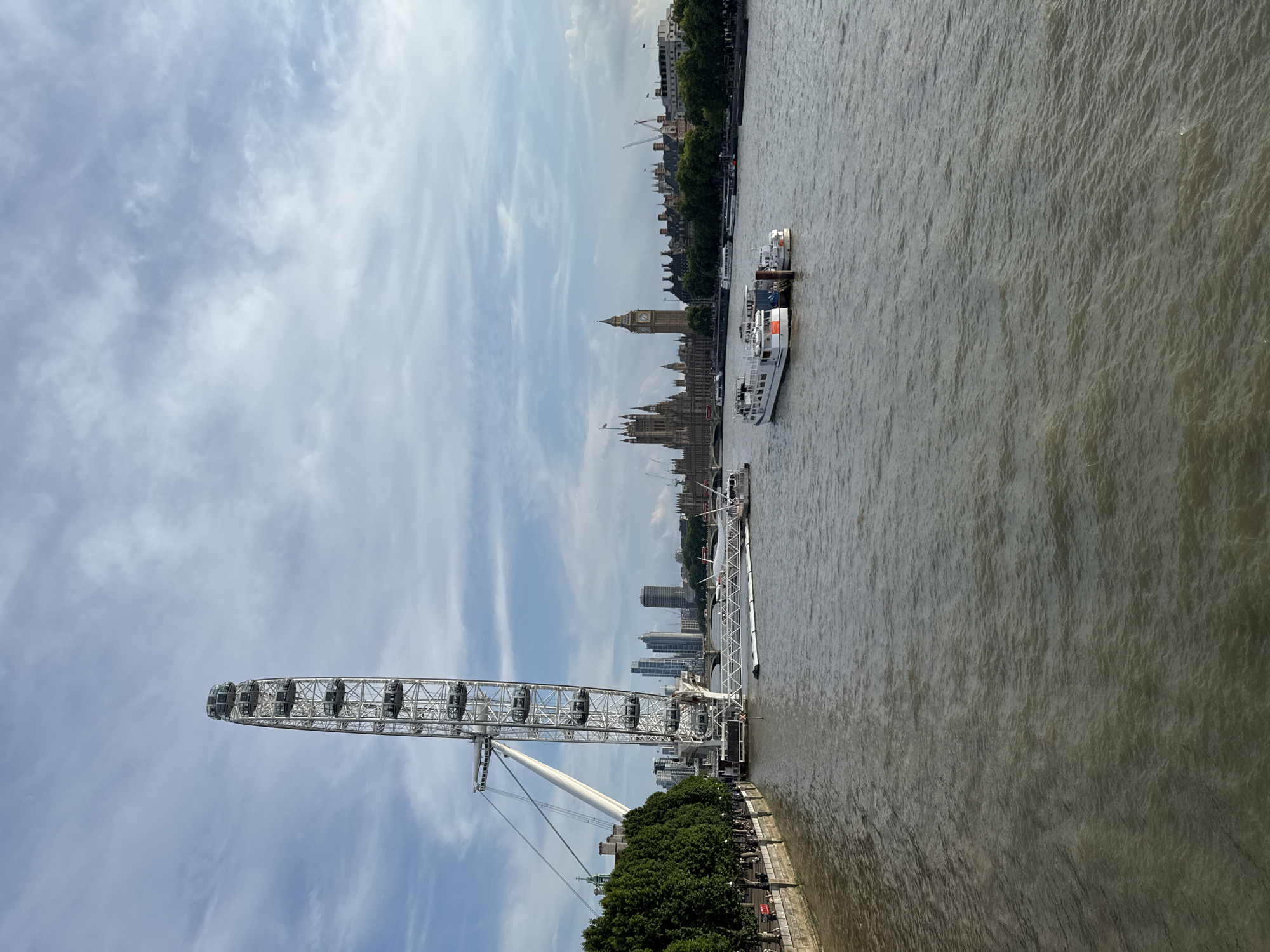 This image captures a scenic view of the River Thames in London, United Kingdom. On the left side of the frame, the iconic London Eye, a large observation wheel, dominates the skyline. Its white structure stands tall against a partly cloudy sky. The riverbank to the left is lined with trees and a promenade with a few people walking.
Across the river, the Houses of Parliament, including the Elizabeth Tower (commonly known as Big Ben), are visible in the distance, showcasing classic Gothic architecture. Several boats are present on the river, including two white passenger boats docked near the center of the image. In the background, more buildings and cranes suggest the ongoing development and urban landscape of London. The water of the Thames appears a murky brown, with ripples indicating a gentle flow. The overall lighting suggests it is daytime, possibly late afternoon or early morning, with soft light illuminating the scene.