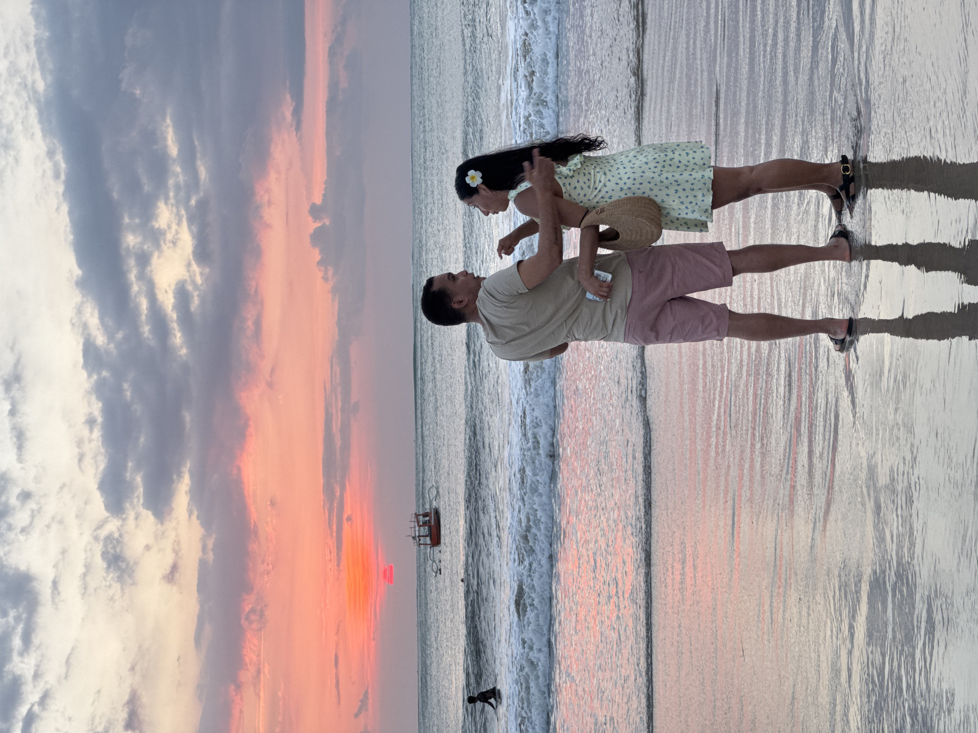 This image depicts a romantic sunset scene on a beach, likely in Kuta Selatan, Indonesia, given the context. The main subjects are a man and a woman, standing in the shallow, reflective ocean water. The man, wearing a light-colored t-shirt and pink shorts, is barefoot. He looks at the woman, who is dressed in a light, patterned dress with a white flower in her dark hair and holding a straw hat. She is leaning towards him, with her hand on his arm, and appears to be in an affectionate pose, looking back at him.
The sky above is a dramatic canvas of sunset colors, transitioning from soft grey and white clouds to vibrant hues of orange and pink near the horizon, where the setting sun is visible. The ocean water is calm, with gentle waves breaking near the shore, and the wet sand and water beautifully reflect the sky's fiery colors. In the mid-ground, a small boat with a red structure can be seen on the water, adding a detail to the tranquil landscape. The overall mood of the scene is peaceful and intimate, capturing a tender moment between the couple against a breathtaking natural backdrop. No visible text is present.
