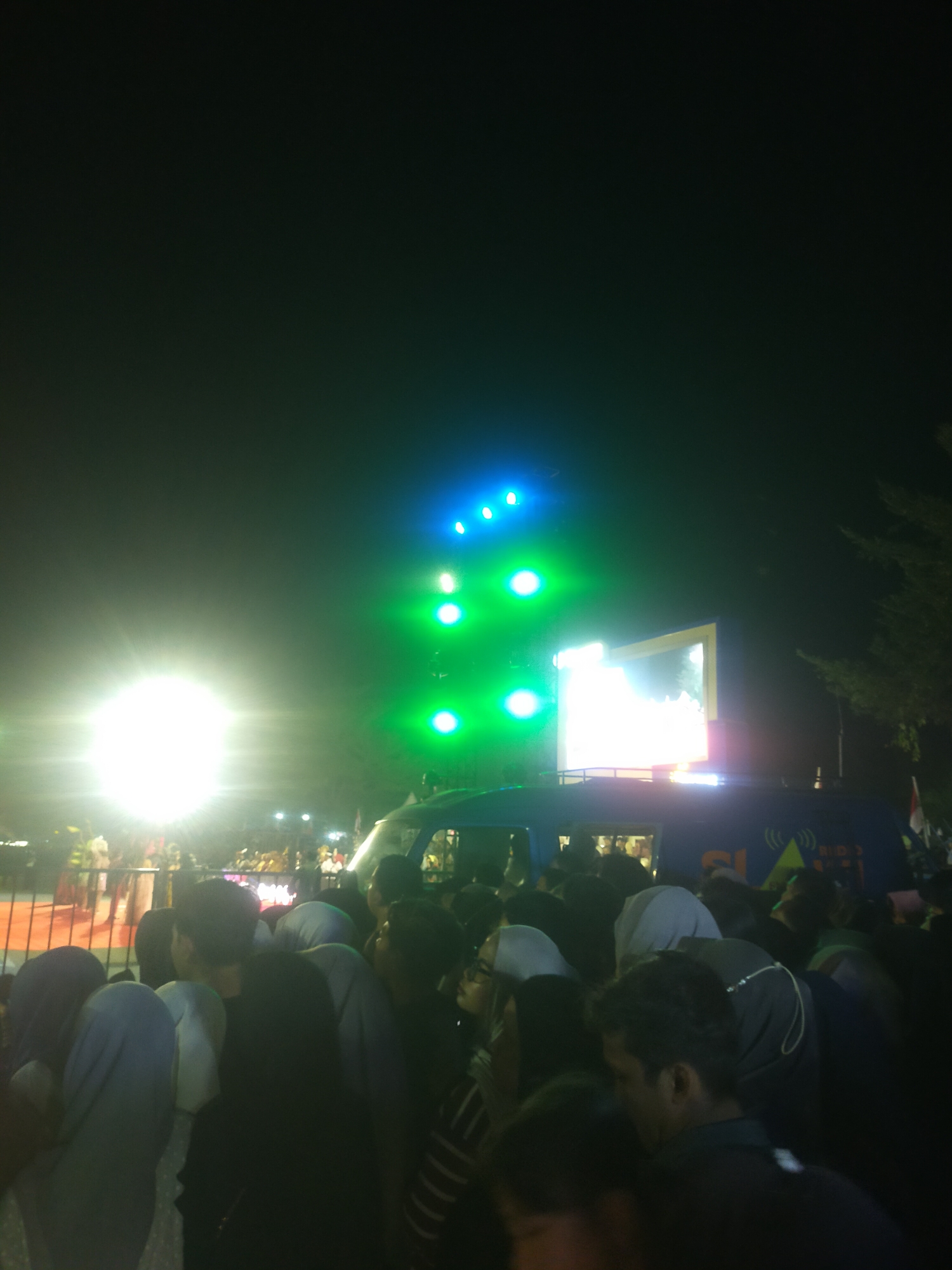 The image depicts a nighttime outdoor event in Slawi, Indonesia, likely a concert or festival, with a large crowd gathered. A bright, white spotlight illuminates the left side of the frame, casting a glow on the surrounding people and a barrier. On the right side, bright green and blue stage lights are visible, along with a large screen mounted on top of a blue vehicle, possibly a mobile stage or broadcasting unit. The vehicle has graphics on its side, including the word "RADIO" and a logo. People are densely packed, many wearing headscarves, and are looking towards the stage or screen. The overall atmosphere is one of excitement and engagement.