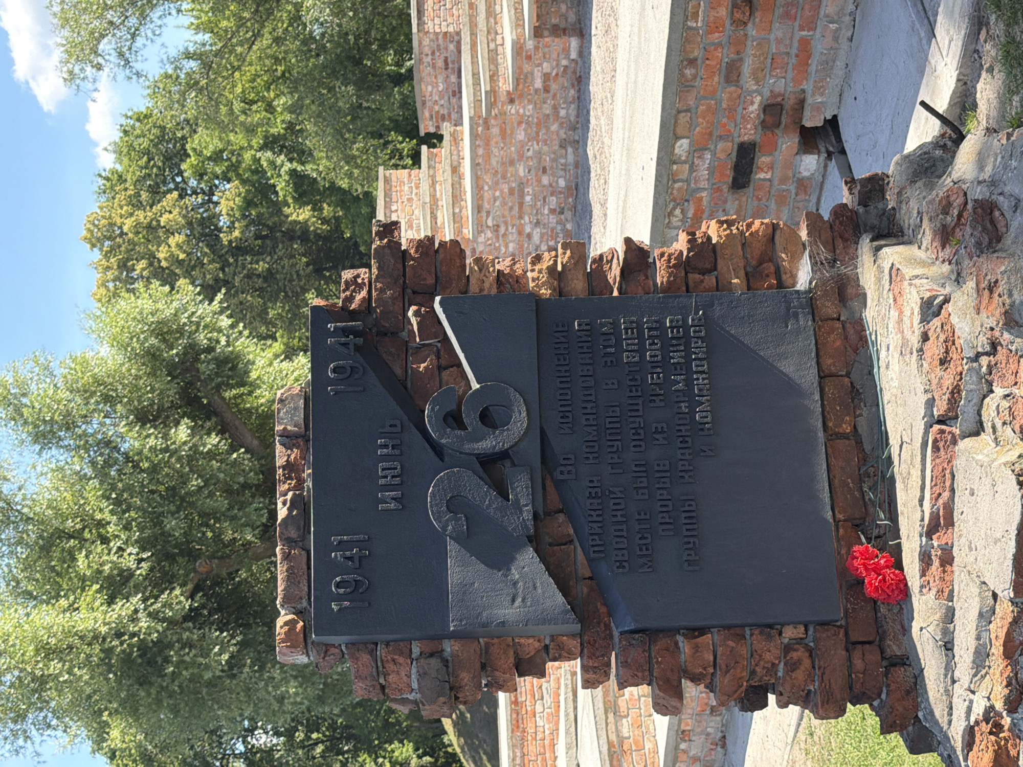 This is a medium, eye-level, outdoor, daytime shot of a memorial monument in a park-like setting. The monument is constructed from dark stone and red brick, with two large, dark stone slabs mounted on a brick base. The left slab is a tall rectangle with the year "1941" etched in its upper portion, followed by the word "ИЮНЬ" (JUNE) below it. To the right of this, a large, cracked numeral "26" is prominently displayed. Below the "26", a smaller dark stone slab contains Russian text. The right side of the monument is a partial structure of exposed red brick. The monument is partially overgrown with green grass and is surrounded by other brick structures that appear to be part of a tiered seating area. Two red carnations are placed on the stone base in the foreground. In the background, mature trees with lush green foliage form a backdrop against a bright blue sky with scattered white clouds. The overall impression is one of solemn remembrance and historical significance.