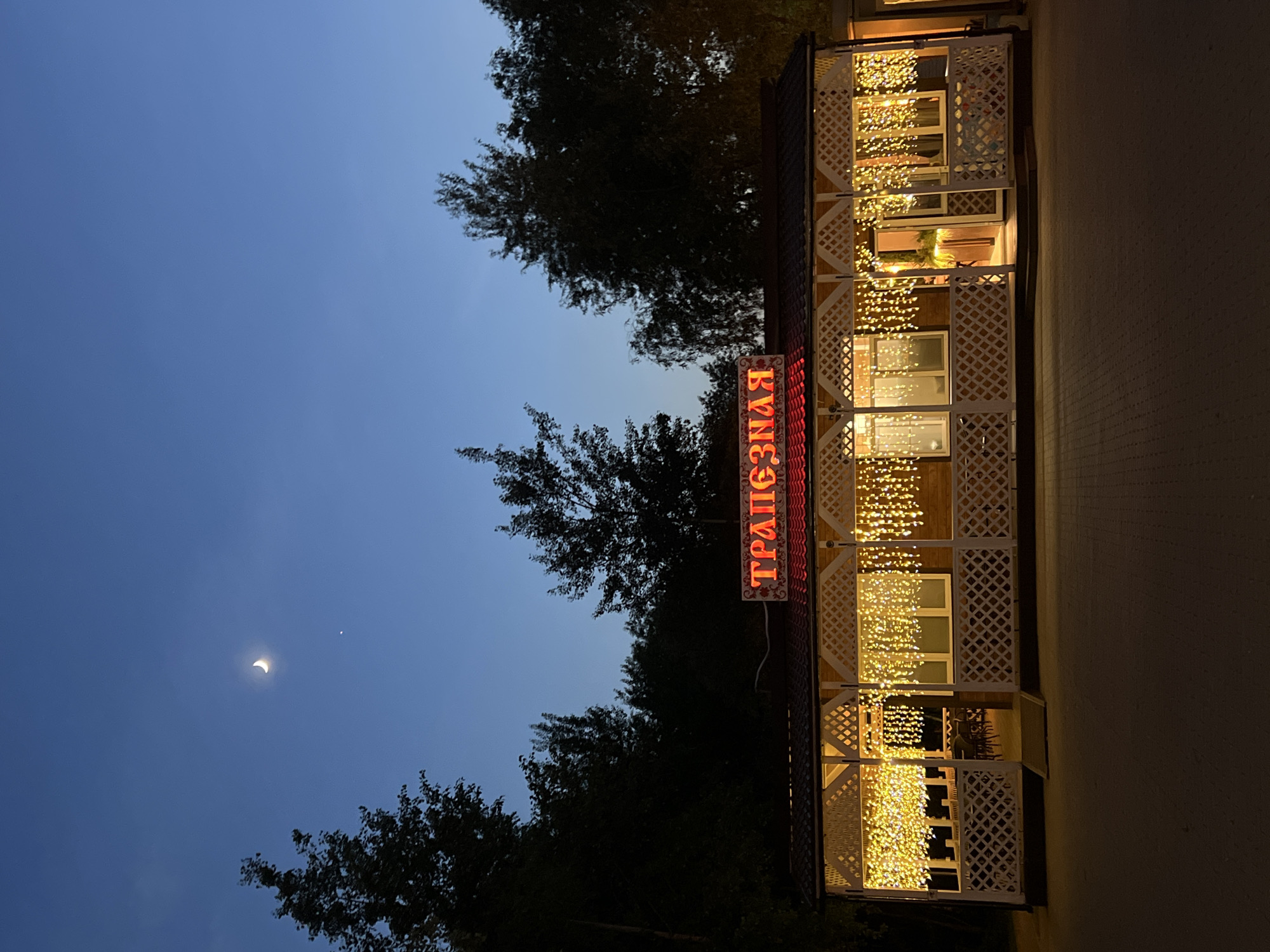 The image shows a three-story building at dusk in Vnukovo, Russia. The building is decorated with lattice and illuminated with numerous strings of small yellow lights. A neon sign in Russian, "ТРАПЕЗНАЯ," is visible on the building. The sky is a gradient of blue, with a crescent moon and a bright star visible. Trees line the left side of the frame. The ground to the right of the building is paved.