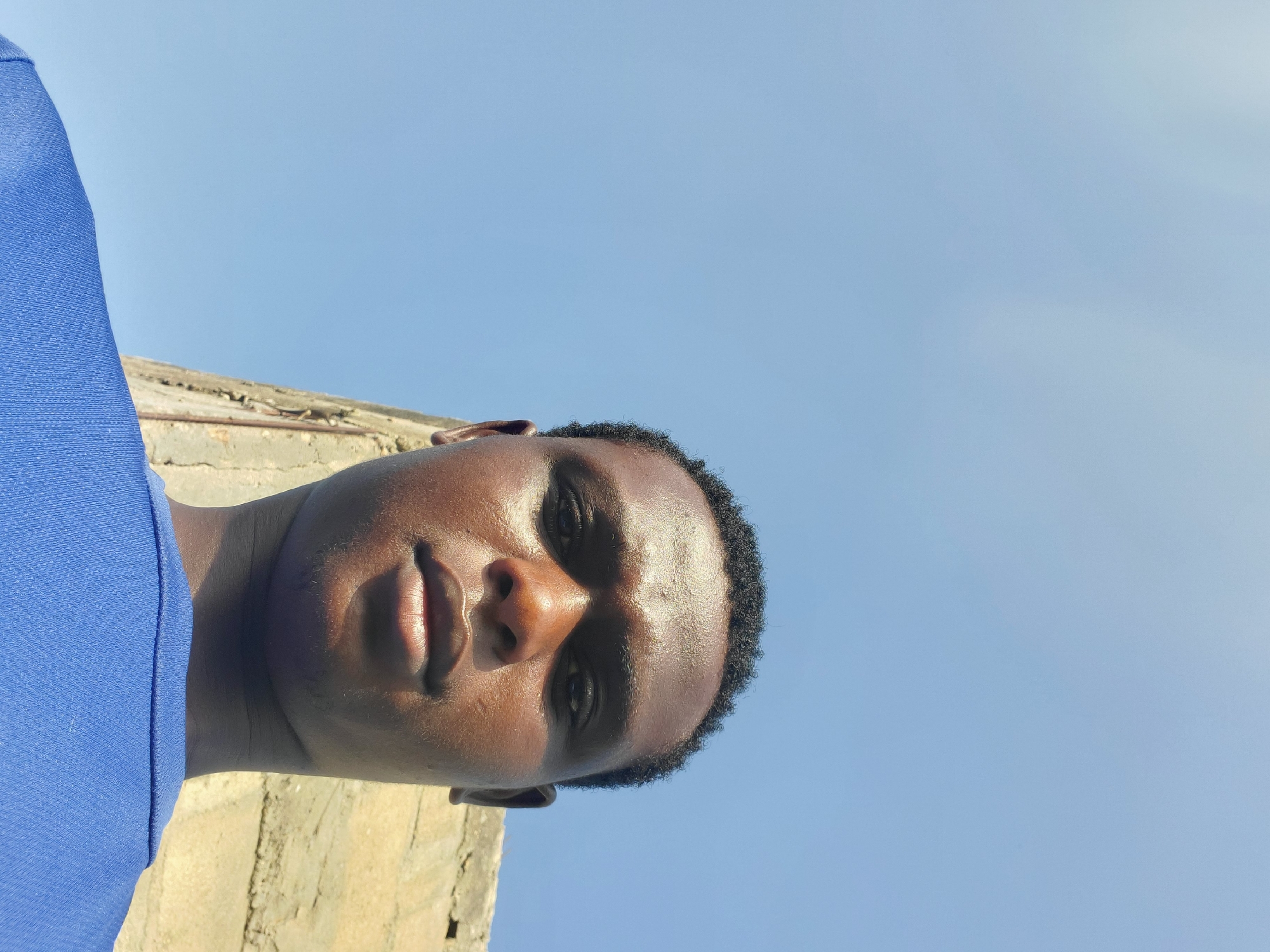 A low-angle, close-up, selfie-style photograph captures a dark-skinned young man against a bright blue sky and a weathered concrete wall, hinting at an outdoor urban setting in Jalingo, Nigeria. The man faces the camera directly, his head tilted slightly upwards. His skin is glistening, suggesting sweat or oil, and the sunlight is strong, creating highlights on his forehead and nose. He wears a bright blue t-shirt, the fabric textured and slightly creased. His hair is short and dark. The background consists primarily of the vast, clear sky, with a portion of a rough, beige concrete structure visible to the right of the man's head. The lighting indicates it is daytime.