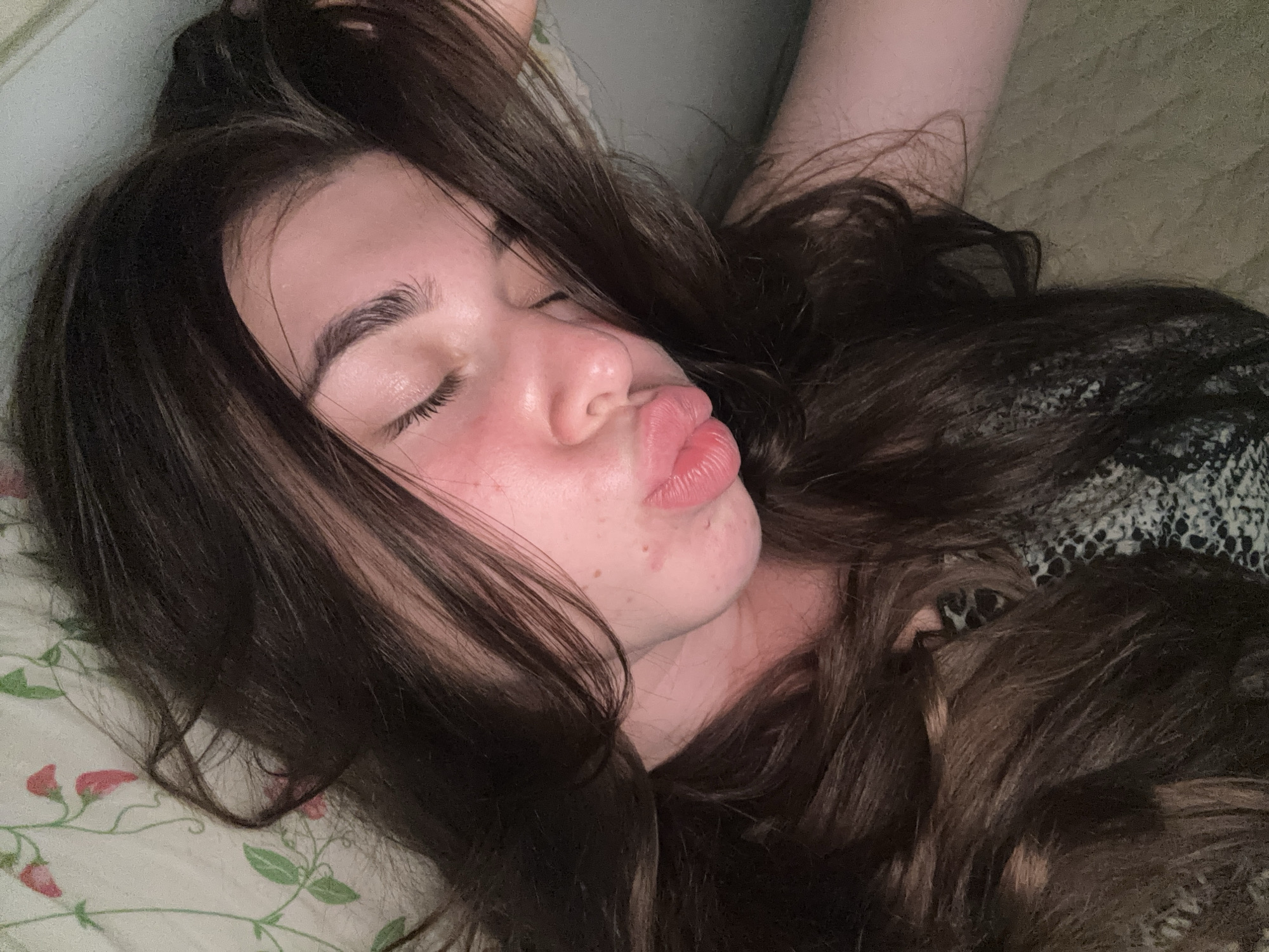 A young woman with long, dark brown hair is lying down, her eyes closed and her lips pursed as if for a kiss. Her face is flushed, and she has a few blemishes. She is wearing a patterned top. The background appears to be a bed with a floral-patterned sheet and a textured blanket. The lighting is soft, suggesting an indoor setting, possibly a bedroom. There is no discernible location in the city of Мещёры, Russia.