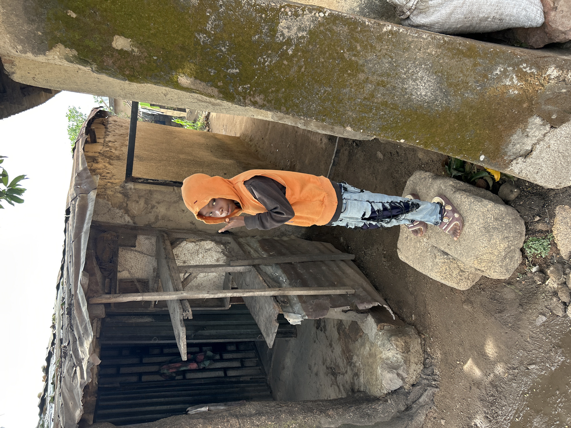 A child stands on rocks between two structures. The child wears an orange hooded sweatshirt, a dark-colored undershirt, light-wash jeans, and sandals. The structure on the left appears to be a shed or small building with a partially collapsed corrugated metal roof and some wooden supports. The structure above is a large concrete or stone barrier covered in moss. The ground is dirt and gravel. The location is Jos, Nigeria.